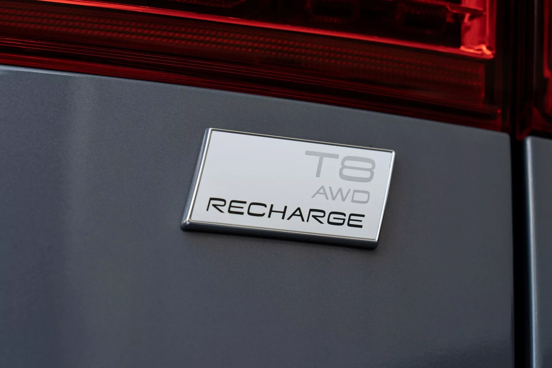 Närbild diagonalt på Volvo XC60 2021 (facelift av andra generationen) bakre emblem. Emblemet läser 'T8 AWD RECHARGE', vilket indikerar en T8-motor, fyrhjulsdrift och ett hybrid- eller elektriskt drivsystem under 'Recharge' varumärket. Positionerat på bakluckan med en del av den bakre lampan synlig, är emblemet på en rektangulär platta med en kromfinish, mot en mörkfärgad, glansig och väl underhållen bilkaross.