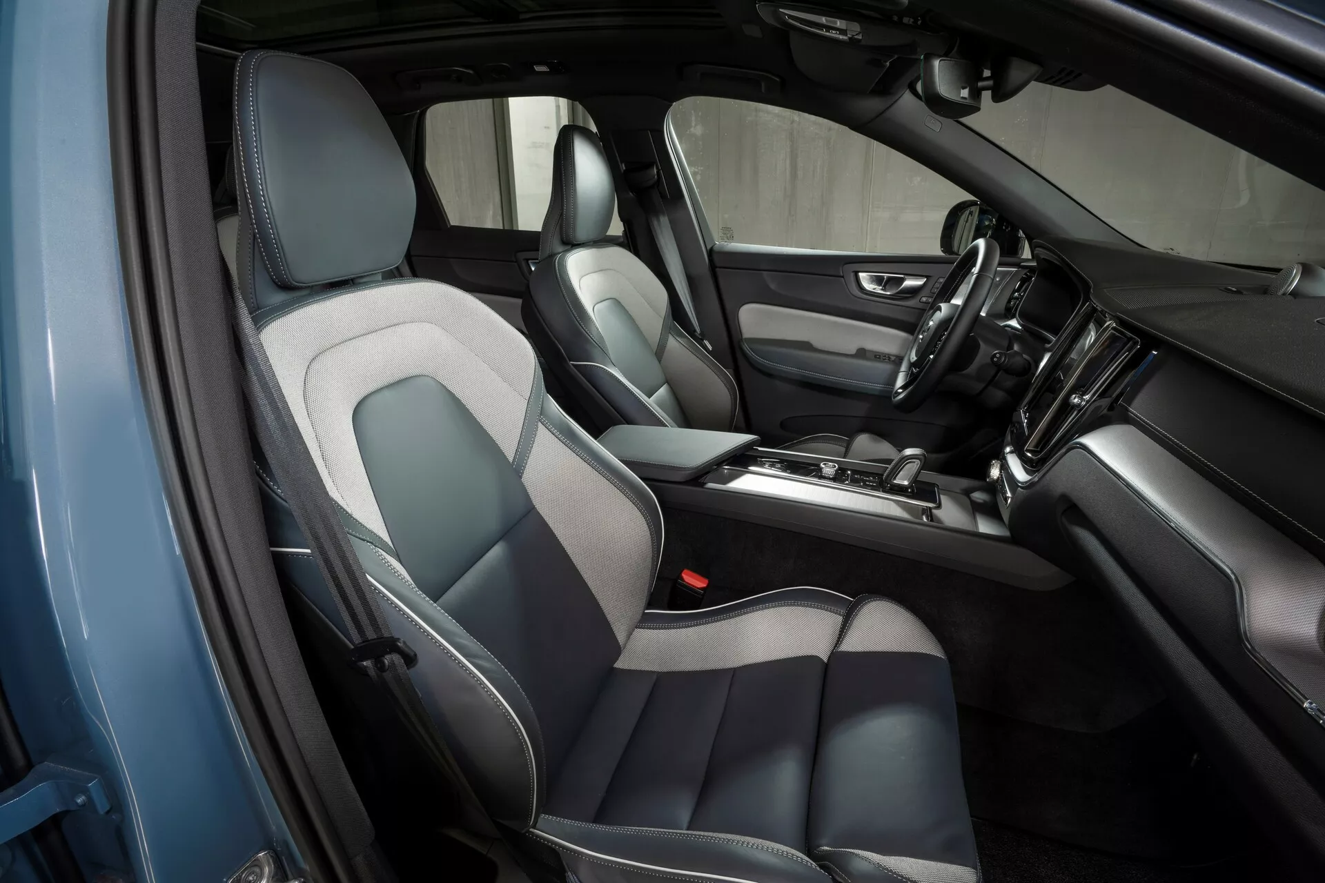 Interiör av Volvo XC60 2021 (2:a generationen-facelift) fotograferad från en upphöjd, sned vinkel från förarsidan, som visar designen och funktionerna i den främre kabinen inklusive säten i läder och tyg i grått och svart, en mittkonsol med växelspak och olika kontroller, en instrumentbräda med en monterad pekskärm för infotainment, kontinuitet i dörrpanelens design och synliga högtalargaller.