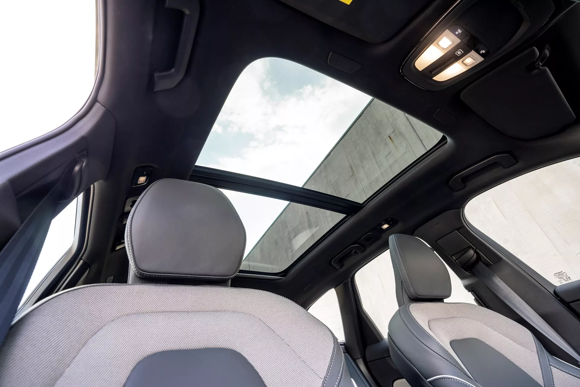 Interiör av Volvo XC60 2021, andra generationens facelift från baksätet, fokuserad på den övre delen av kupén. Anmärkningsvärda funktioner inkluderar ett stort panoramatak med solskydd, svart innertak som indikerar en högre utrustningsnivå eller lyxpaket och kabinbelysning i taket. Bilden fångar en del av nackstödet och den övre ryggen på framsätet samt B-stolpen och handtaget ovanför bakdörren.