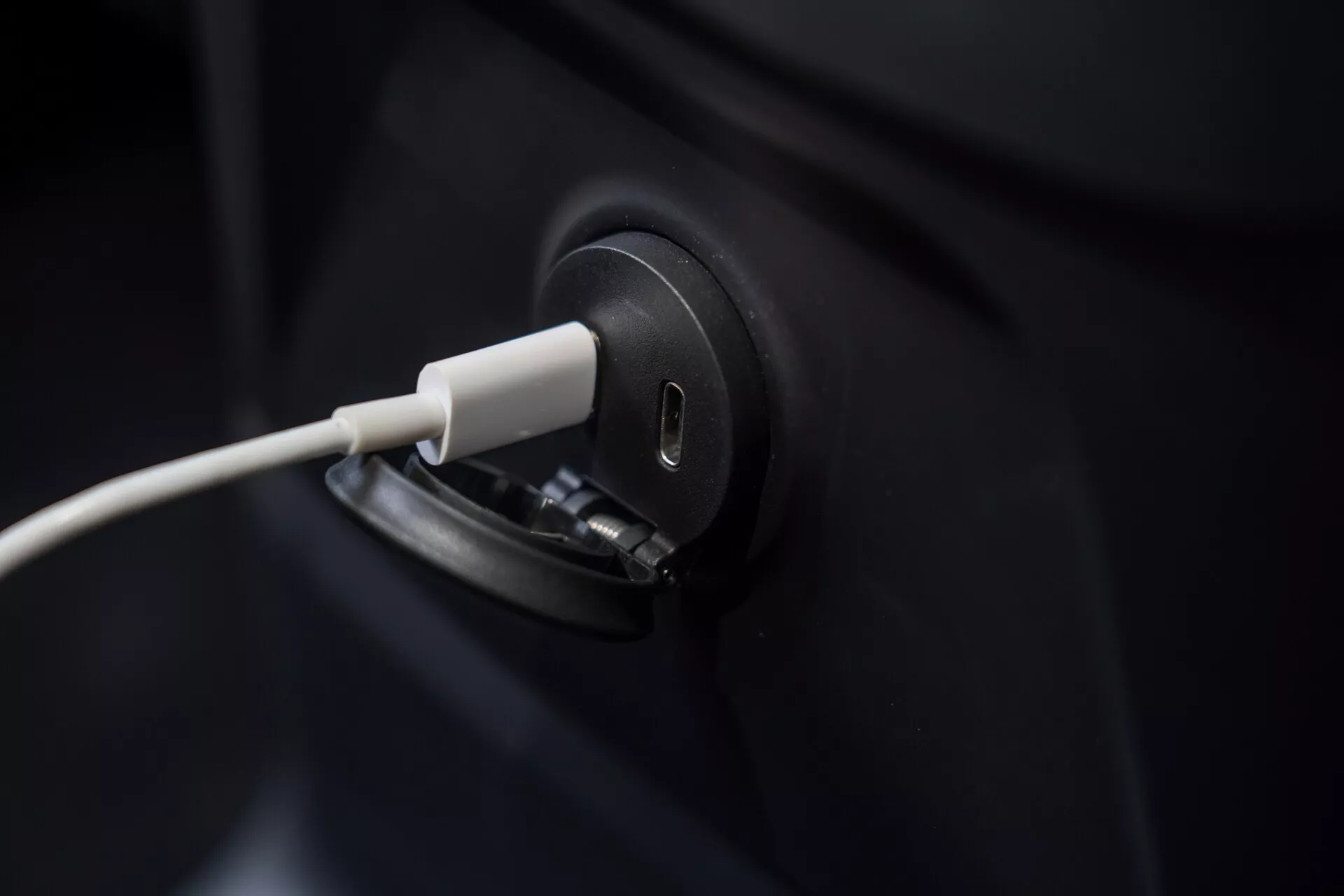 Närbild av laddningsporten på en Volvo XC60 2021 elbil med inkopplat laddkabel, som betonar den snygga och moderna designen. Bilden är tätt centrerad på laddningsporten, vilket lämnar bilens mörka kaross mestadels ur fokus. Den suddiga bakgrunden framhäver precisionen i laddningsprocessen för elbilar, vilket symboliserar grön teknik och hållbar transport.