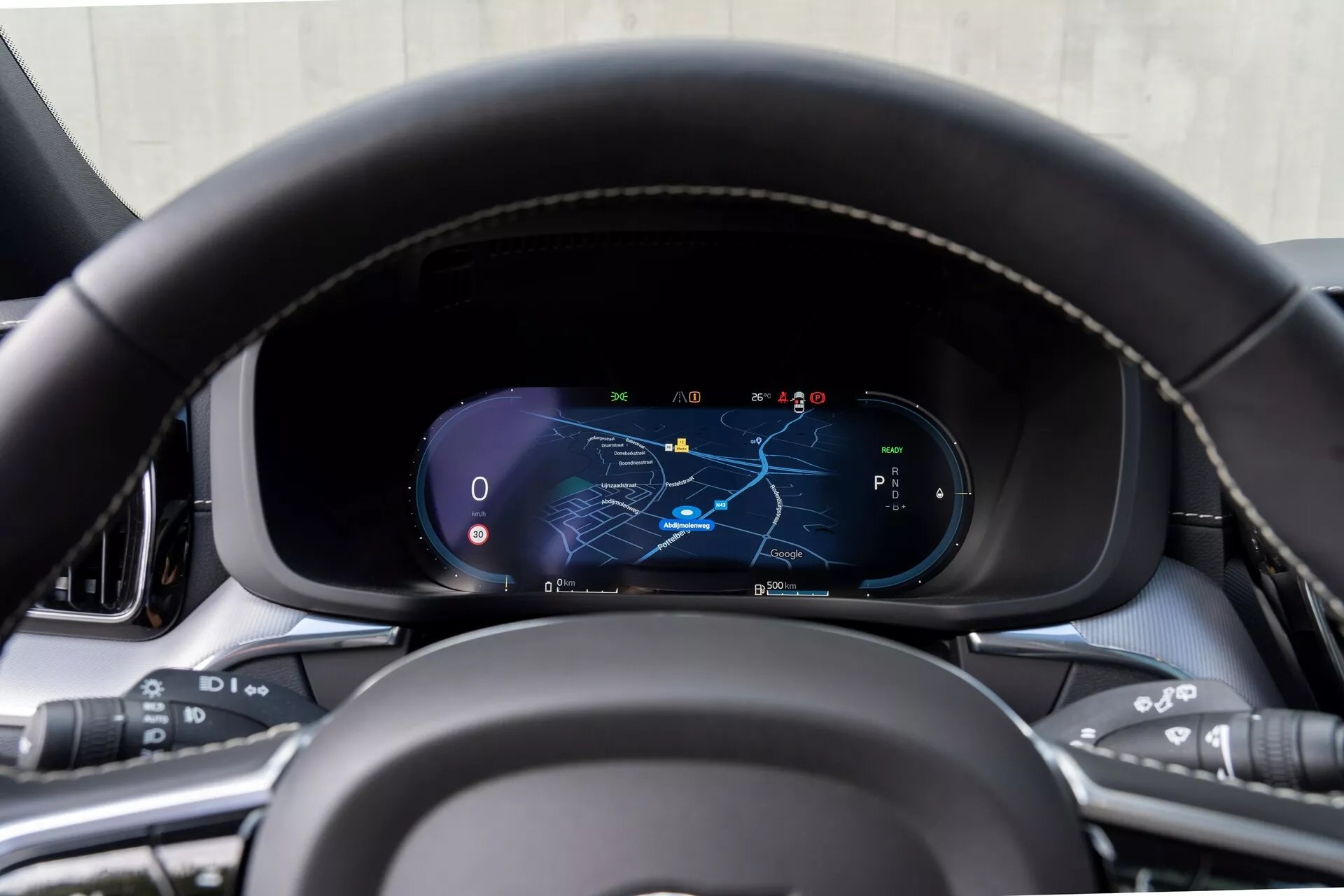 Närbild av instrumentbrädan i Volvo XC60 2021 (2:a generationens facelift) från förarsätets perspektiv, med fokus på den digitala instrumentpanelen och en läderklädd ratt med sydda detaljer och multifunktionsknappar för media, röstkommandon, farthållare och körassistanssystem. Den digitala skärmen visar en karta med markerad navigeringsrutt, hastighetsmätare, varvräknare och växelväljare i parkeringsläge, omgiven av material av premiumkvalitet.