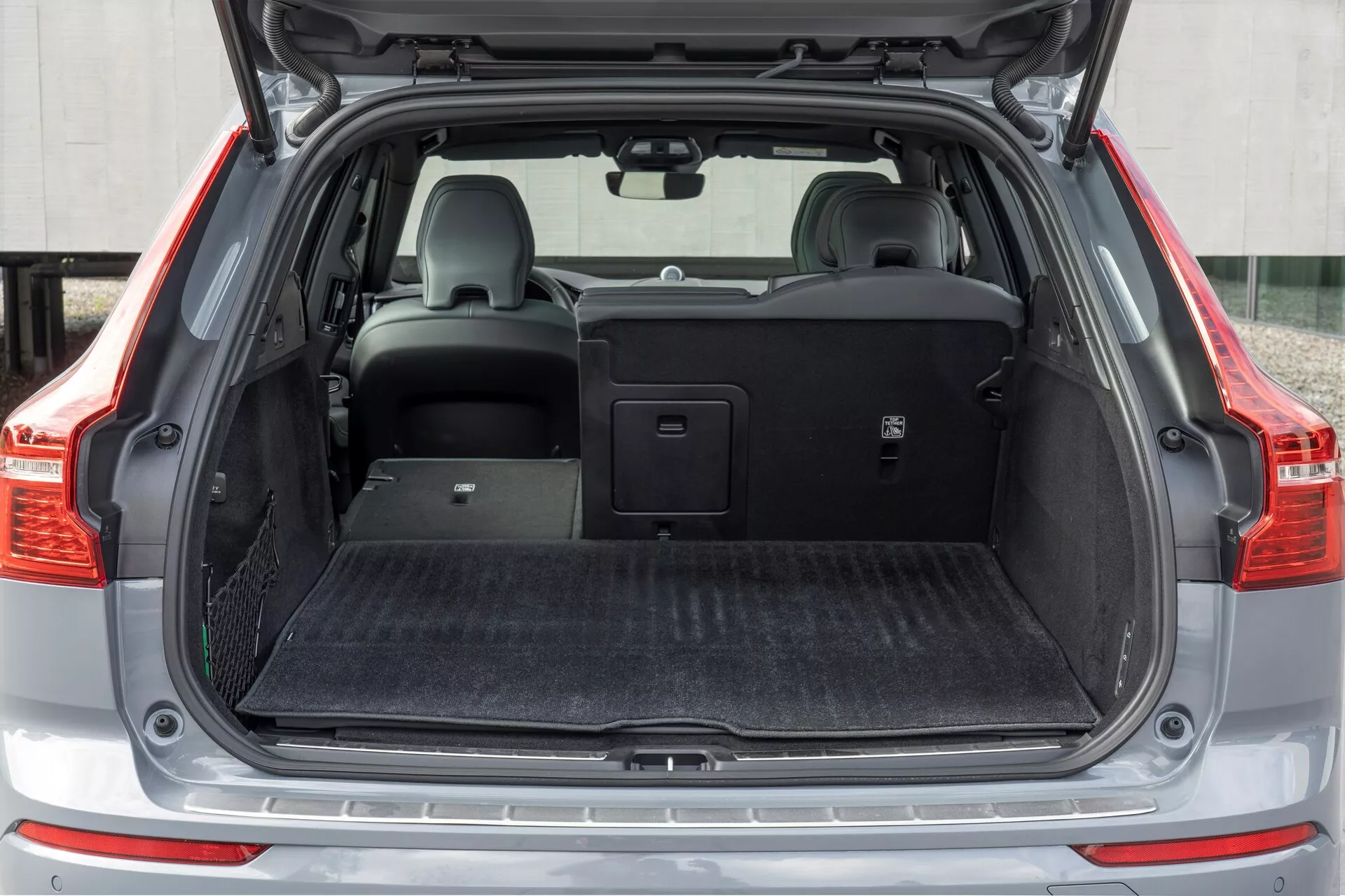 Bilden visar lastutrymmet bak i en Volvo XC60 2021 (ansiktslyftning av andra generationen), sedd från en rak vinkel med den öppna bakluckan. Mellanavståndsbilden fångar hela bredden av lastutrymmet, som har ett plant lastgolv täckt med svart mattliknande material. På vänster sida finns ett litet förvaringsfack med nät. Det högra baksätet är nedfällt och visar den delbara fällfunktionen, medan det vänstra sätet står upprätt med ett nackstöd. Ett utdragbart lastskydd är placerat ovanför de bakre sätena, vilket visar fordonets förvaringsmångsidighet.
