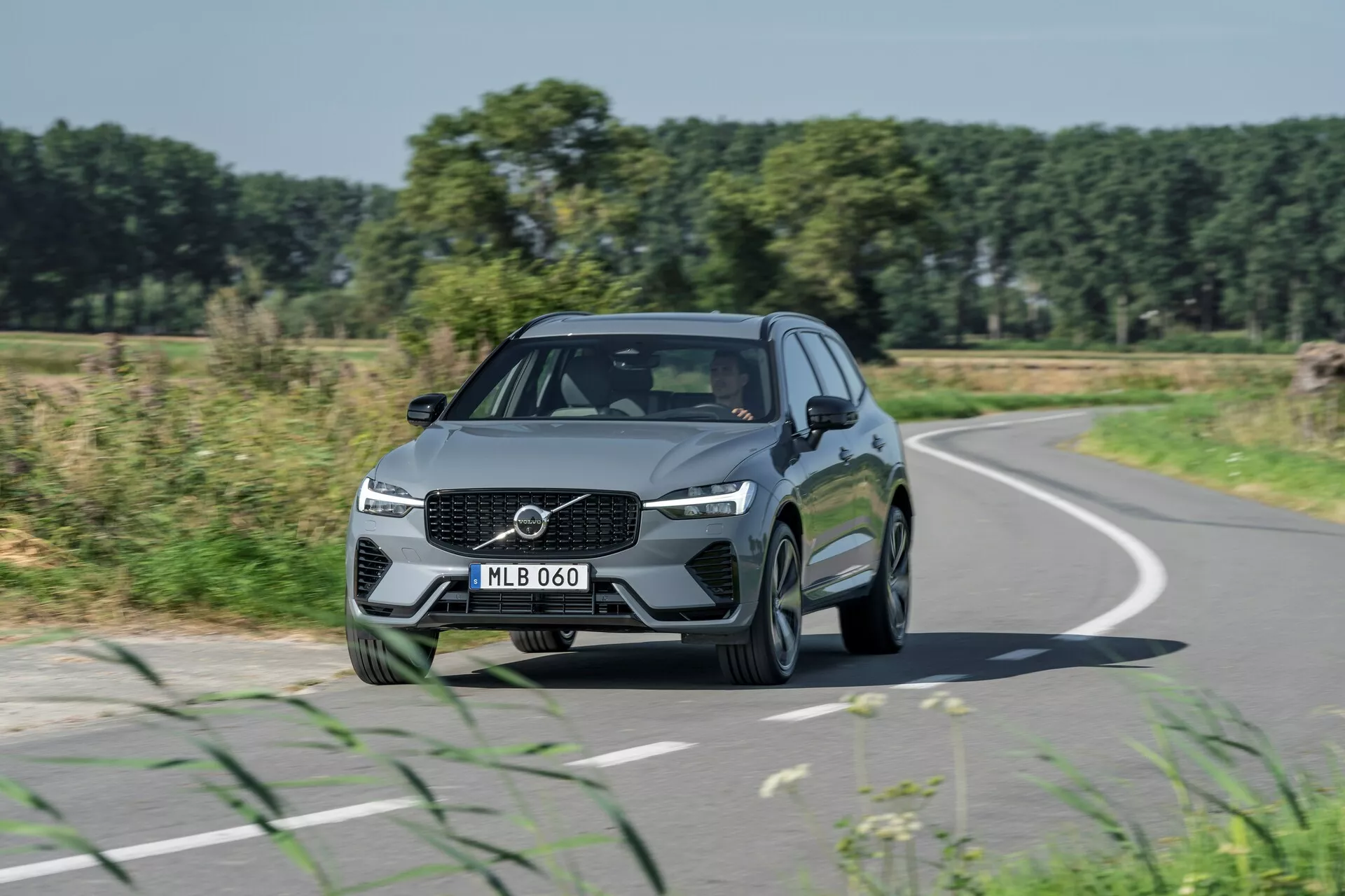 Bilden fångar en Volvo XC60 2021 (2:a generationens facelift) i rörelse från en främre trekvarts-vinkel, och framhäver designelement som en framträdande grill med centralt placerad logotyp, kantiga LED-strålkastare och slanka linjer på huven. Bilen, som färdas på en något krökt väg, har en metallisk nyans och fler-ekrade lättmetallfälgar, omgiven av konturerade linjer och integrerade sidobackspeglar med blinkers. Mot en bakgrund av fält, träd och en klarblå himmel framhäver denna representation bilens design, vägresa och verkliga körscenarier.