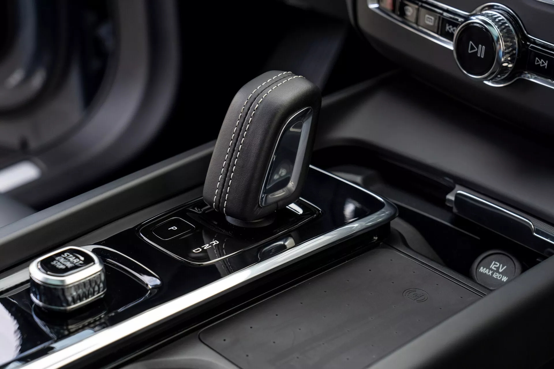 Närbild av mittkonsolen i en Volvo XC60 2021 (2:a generationen, facelift). Bilden framhäver växelväljaren med en ovandel klädd i läder med söm-detaljer, placerad på en blank svart yta. Till höger finns en ratt med räfflad kant, troligtvis för infotainmentsystemets navigering, med en tryckknapp för valfunktion. Bredvid växelväljaren finns knappar för parkeringssensorer och ett automatiskt start/stopp-system. Mittkonsolen har metalldetaljer och en upplyst infotainmentskärm synlig i den suddiga bakgrunden, vilket betonar fordonets moderna, högkvalitativa design och hantverk.