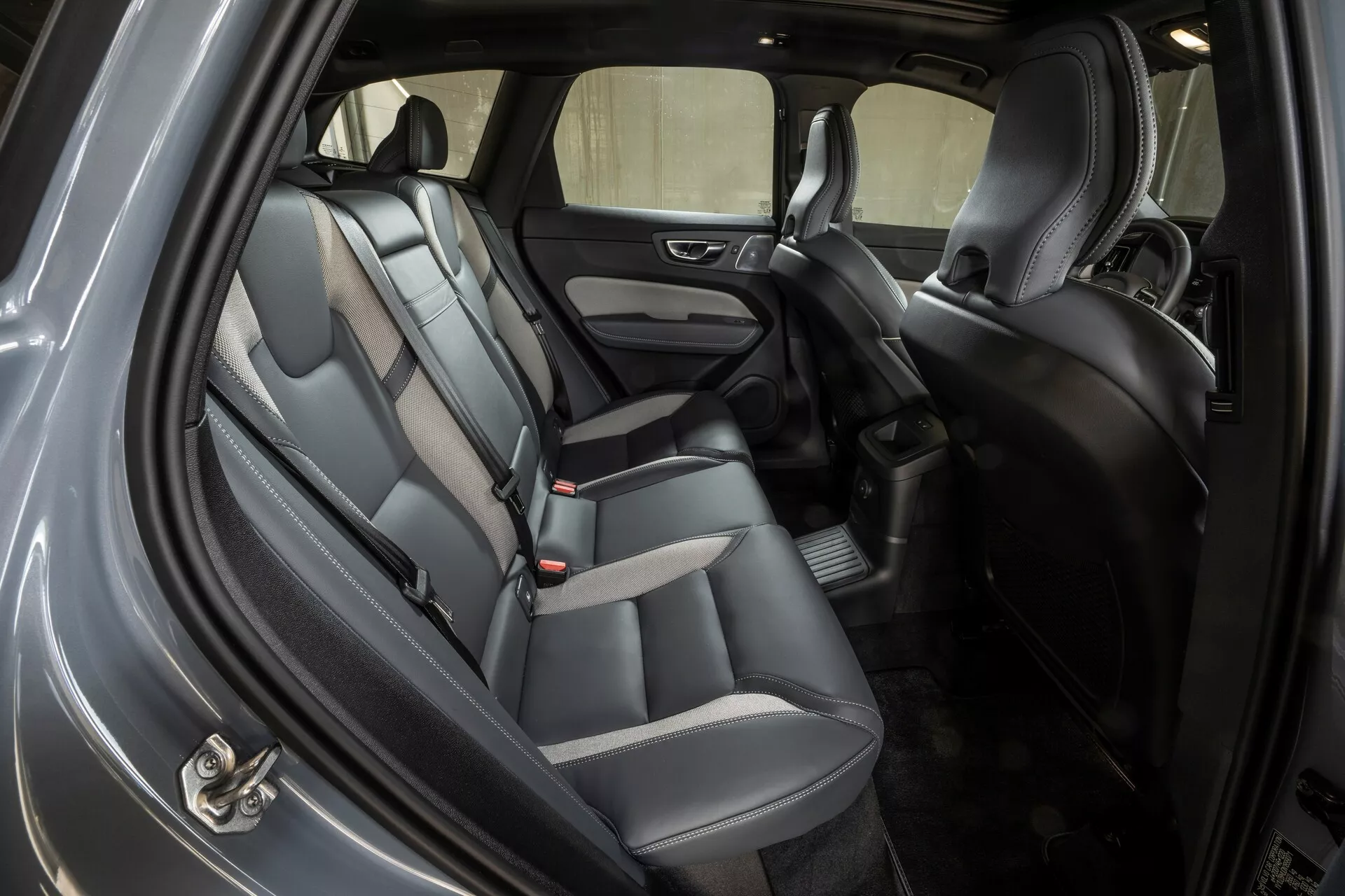 Interiörbild av 2021 Volvo XC60 som visar baksätesområdet från en sned vinkel med baksidanvisardörren öppen. Bilen har tvåfärgade skinn- eller konstlädersäten, nackstöd, säkerhetsbältesspännen för tre passagerare och klädda dörrpaneler med inbyggd högtalare och fönsterhissar. Golvet är täckt med en matta och ett golvunderlag, och mittkonsolen synlig mellan framsätena innehåller förvaringsutrymme. Den väl upplysta kupén antyder omgivande och takmonterad belysning och erbjuder god sikt genom sido- och bakrutorna.