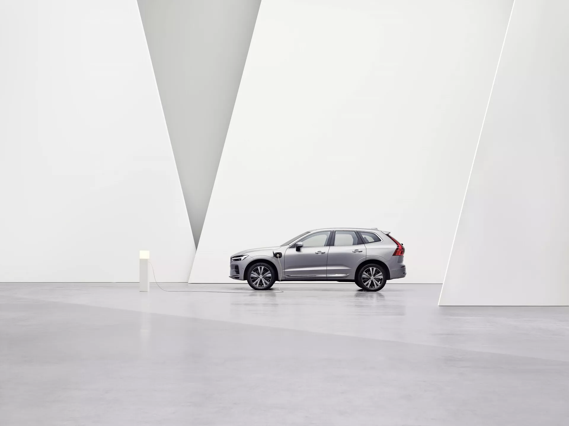 Bilden visar en Volvo XC60 2021 med andra generationens facelift SUV i ett rymligt, minimalistiskt konstgalleri med vita väggar och ett blankt golv. Fordonet, sett från vänster sida, framhäver legeringshjul, karaktärslinjer över sidodörrarna, sidospeglar, tonade bakrutor och distinkta takräcken. Från en lätt låg vinkel är frontgrillen, LED-strålkastarna, integrerade blinkers och parkeringssensorer på främre stötfångaren synliga. Den rena miljön och väggens V-formade partition framhäver bilens design och premiumposition på marknaden under mjuk, diffus belysning.