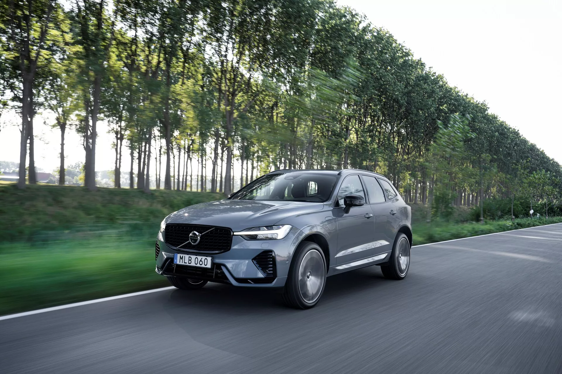 Bilden visar en rörlig Volvo XC60 2021 (2:a generationens facelift) fångad från en trekvarts framvinkel, som visar förarsidan av bilen. Denna moderna lyxiga SUV har en stilren design och sitt emblem synligt på frontgrillen. Tagen med en lätt nedåtgående vinkel inkluderar bilden motorhuven, stötfångaren, grillen, strålkastarna och hjulen. Den mörka metalliska bilen, eventuellt grå eller svart, med en glansig finish, har lättmetallfälgar, LED-strålkastare och en aerodynamisk kaross. Den suddiga bakgrunden av en stadsmiljö med träd antyder rörelse och betonar bilens prestandaegenskaper.