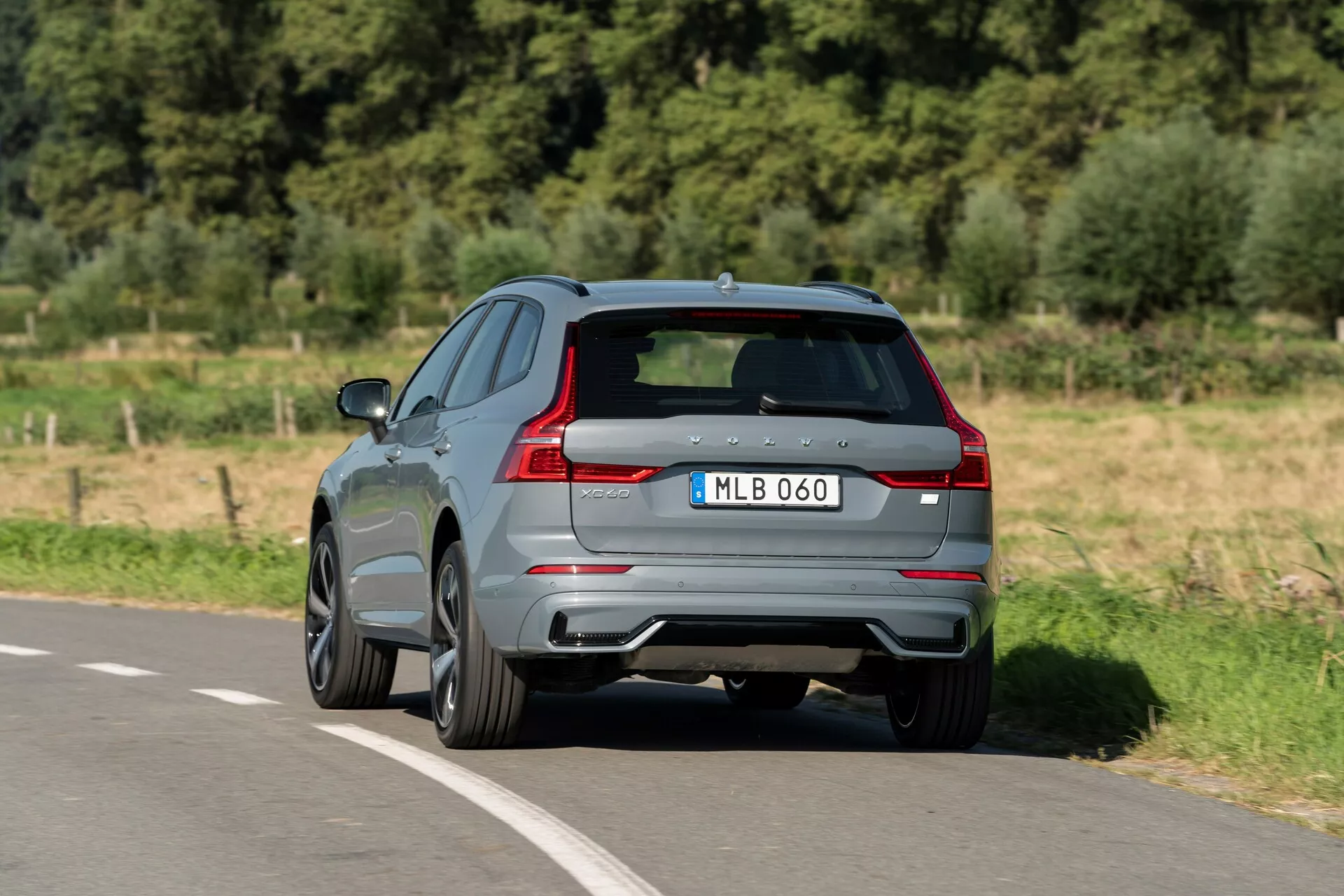 Trekvarts bakifrån vy av en 2021 Volvo XC60 som rör sig längs en asfalterad väg. Fotograferad från en upphöjd vinkel visar denna moderna SUV sin gråa glansiga färg, distinkta LED-baklys, dubbla avgasrör och bakre diffusor. Tillverkarens emblem och modellbeteckning är synliga på den skulpterade bakluckan, kompletterade med tonade rutor och lättmetallfälgar med flerspårigt mönster, vilket antyder en sportigare variant. Bilden ger en klar inblick i den aerodynamiska designen och högkvalitativa funktioner som är kännetecknande för ett modernt fordon.