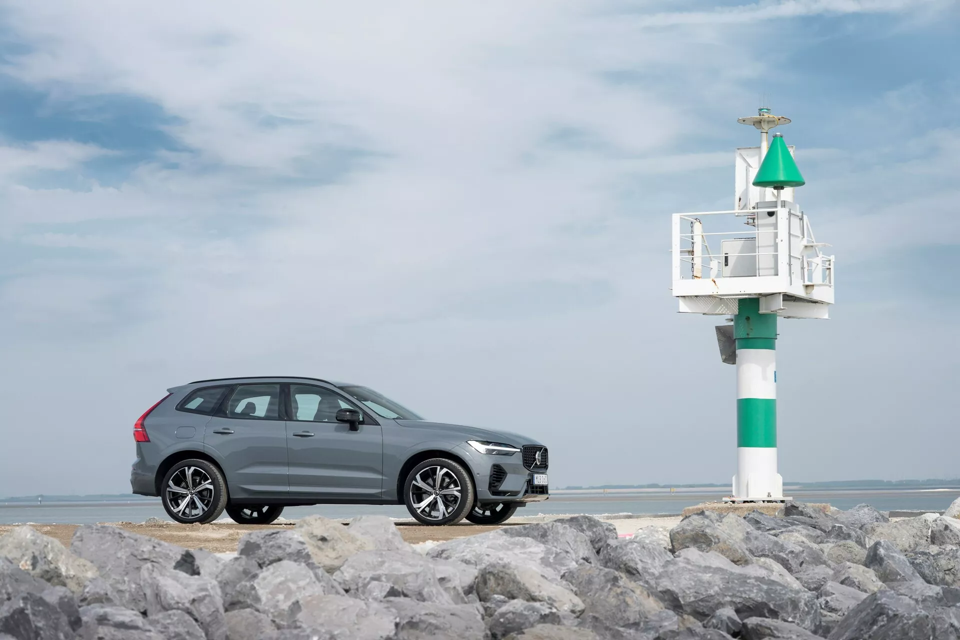 Profilvy av en grå 2021 Volvo XC60 (andra generationen, facelift) SUV på stenig mark med ett maritimt sjömärke i bakgrunden. Den eleganta designen inkluderar kromfönsterlister, multi-ekrade aluminiumfälgar och integrerad blinkers i sidospegeln, fångad mot en delvis molnig kusthimmel.