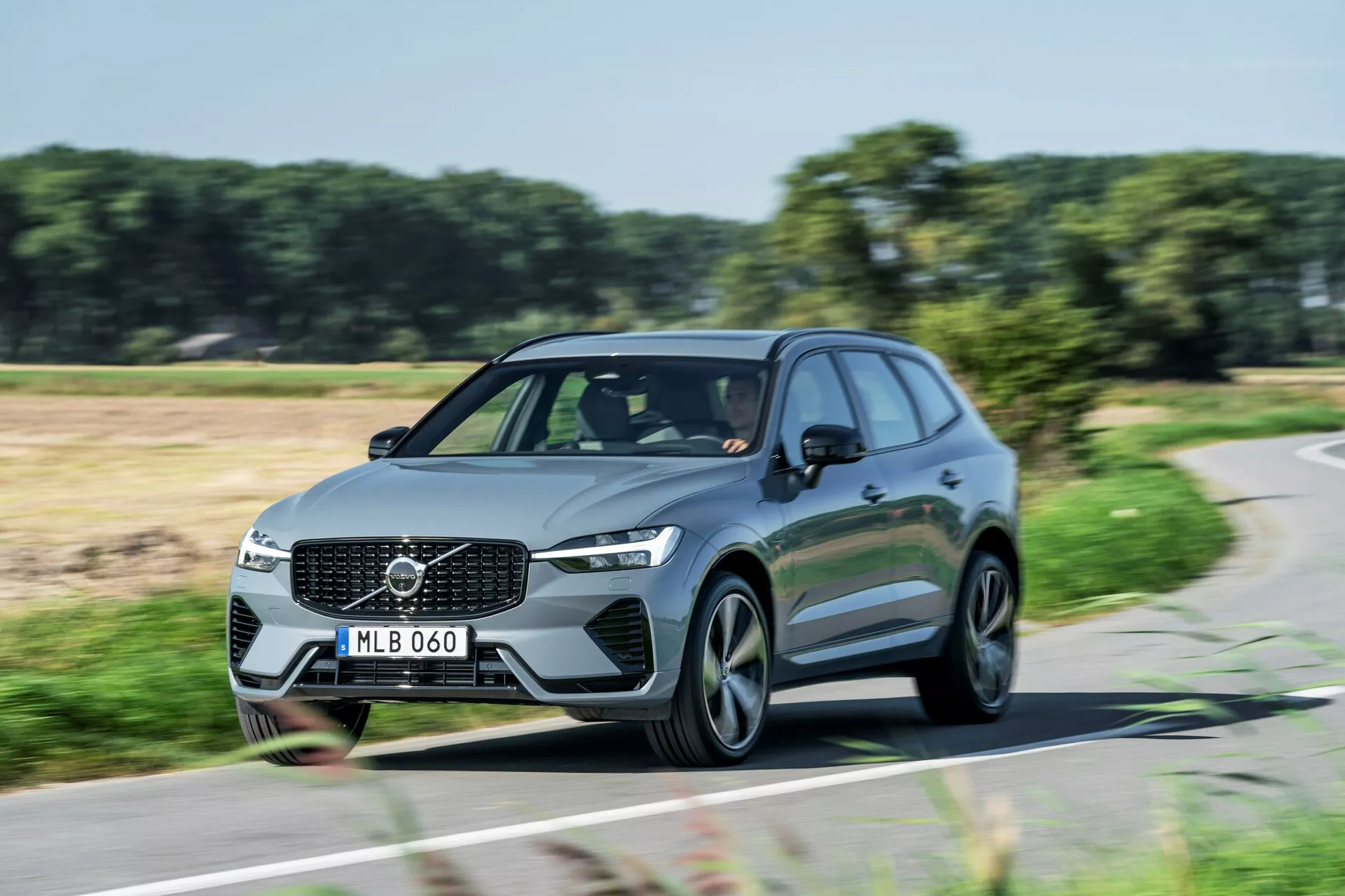 Dynamisk dagsljusbild av en rörlig Volvo XC60 2021 (andra generationens facelift) SUV fotograferad från en trekvarts framvinkel. Bilden visar frontgrillen med en framträdande Volvo-emblem, vinklade LED-strålkastare, aerodynamiska linjer och moderna fler-eker fälgar i aluminium. De tända dagsljuslamporna indikerar att bilen är i rörelse. Fotot framhäver den eleganta designen och väg närvaro av detta moderna familje- eller lyxfordon.