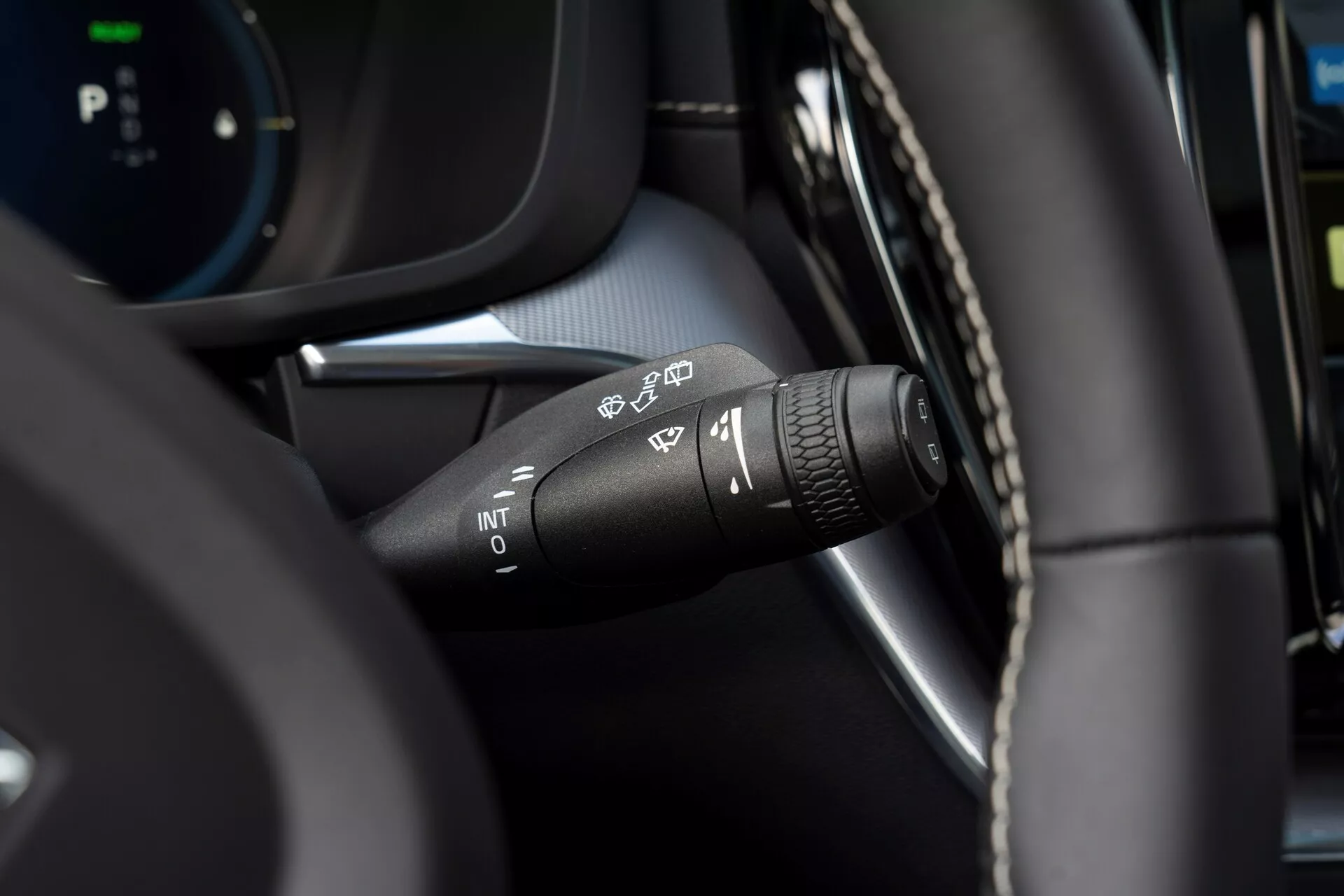Närbild av en vindrutetorkarkontroller i en Volvo XC60 2021 (andra generationen facelift) från förarens perspektiv. Fokus på olika torkarsymboler: automatisk regnsensor, intermittent, låg hastighet och hög hastighet. På spakens spets finns en roterande ring för att justera intervallet för intermittent torkning. Modern interiör med en blandning av läderliknande material och svarta plastkomponenter, med en suddig digital instrumentpanel i bakgrunden.