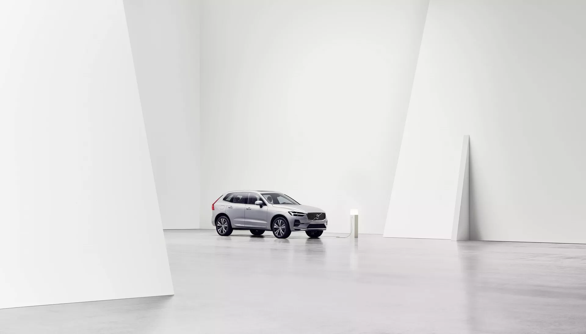 Bild av en Volvo XC60 2021 (2:a generationen, facelift) placerad centralt i en minimalistisk, rymlig och välbelyst studiomiljö, tagen från ett trekvarts främre perspektiv. Bilens ljusa silverfärg kontrasterar med de mörkare hjulen och en del av den främre grillen, och visar moderna SUV-designelement som böjda och vinklade linjer. Utsikten framhäver den främre grillen, strålkastarna, motorhuven, vindrutan och förarsidan, inklusive fram- och bakdörrar samt sidorutor. Den rena vita bakgrunden framhäver bilen, fokuserar uppmärksamheten på dess design och egenskaper, lämplig för marknadsförings- eller konstnärliga ändamål.