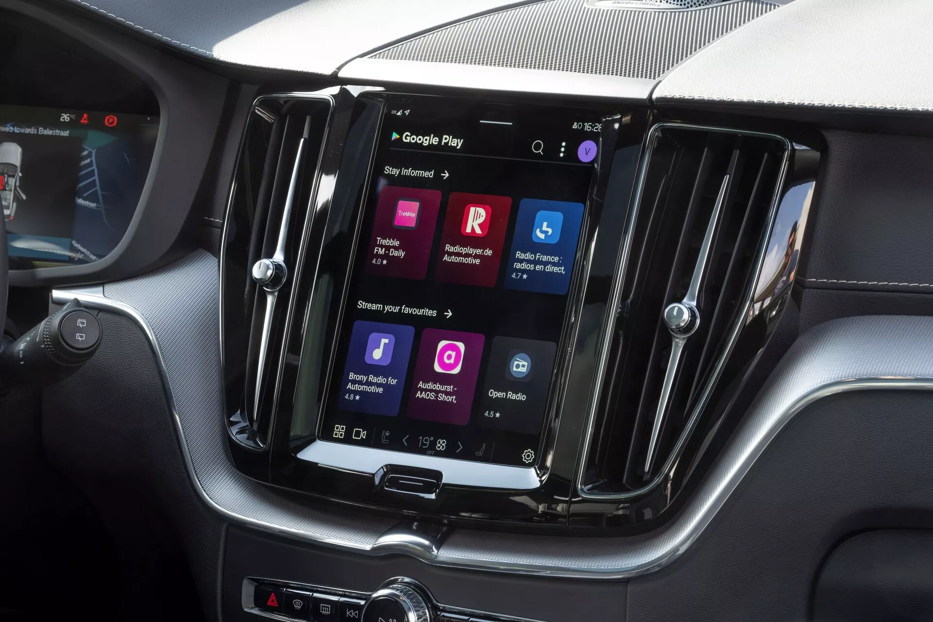 Närbild av Volvo XC60 2021:s centralkonsol med ett toppmodernt vertikalt orienterat pekskärms-infotainmentsystem. Skärmen är inramad i en glansig, premium-finish med silverdetaljer. Skärmen visar färgglada ikoner, inklusive Google Play, vilket indikerar avancerad programvara med appanslutning, röstigenkänning, internetåtkomst och multimediaströmning. Snygg, modern design med kolfibertexturer på instrumentpanelen. Fysiska knappar för klimatkontroll nedanför skärmen. Bild tagen från framsätesperspektivet, fokuserad på mittsektionen av konsolen, antyder en lyxig bil.