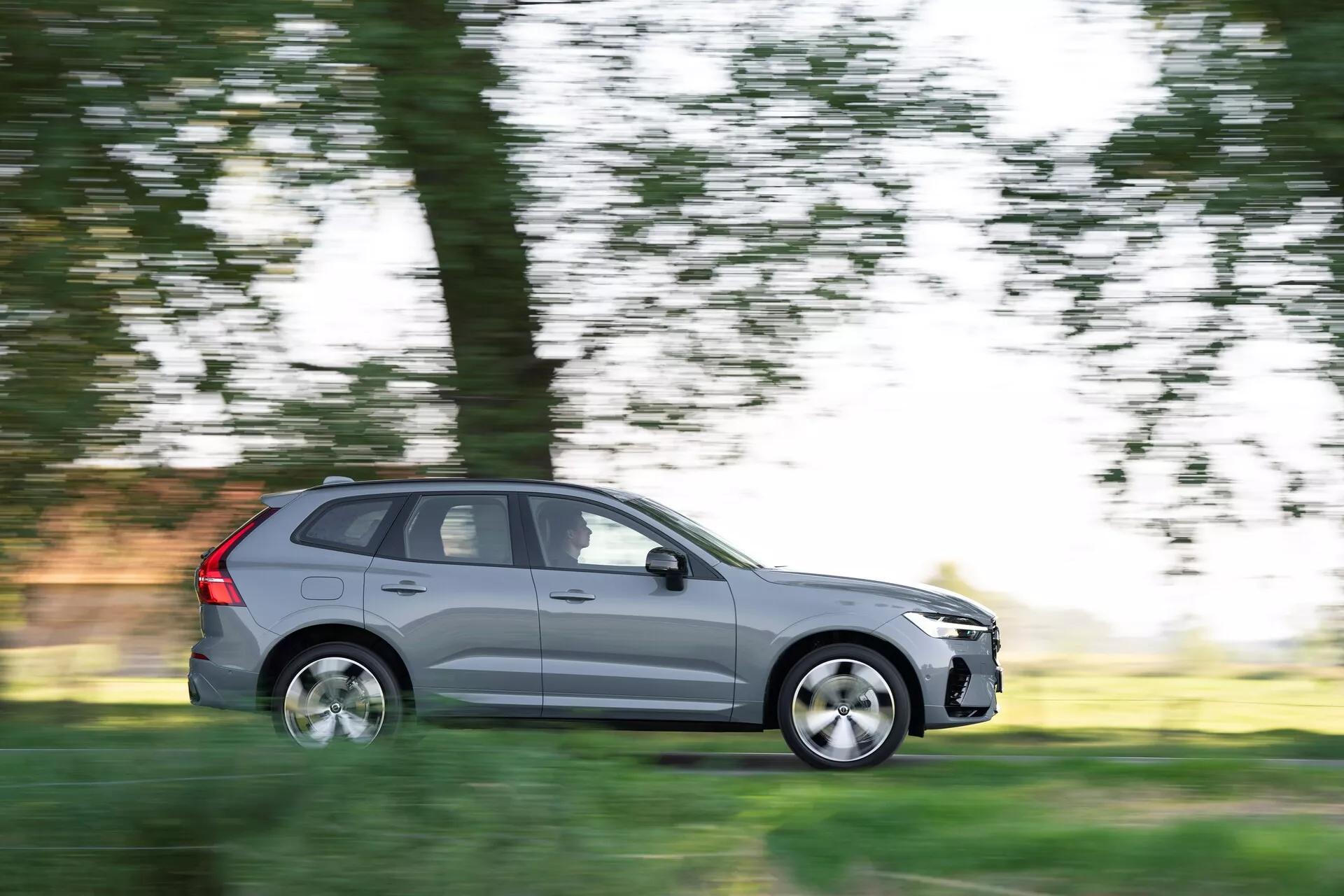 Bilden visar en Volvo XC60 2021 (2:a generationens, facelift) SUV i rörelse, fångad med panoreringsteknik som suddar bakgrunden medan bilen är i skarpt fokus. Sedd från förarsidan i komplett sidoprofil, har den metalliska grå bilen fem dörrar, aluminiumfälgar och standardvägsdäck. Scenen antyder dagskörning genom en naturlig miljö, med suddiga träd och gräs i bakgrunden, vilket betonar bilens rörelse.