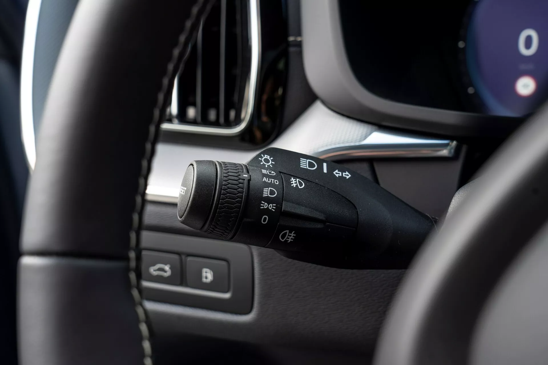 Närbild av interiören i en Volvo XC60 2021 (andra generationens facelift), med fokus på vänstra sidan av rattstången. Huvudämnet är strömställaren för strålkastare och indikatorer med symboler för automatiska ljus, parkeringsljus, strålkastare, dimljusinställningar och en anpassningsbar funktion. Bilden är tagen från förarsätet, något uppåt och till vänster, och visar den suddiga vänstra övre sidan av ratten i förgrunden och betonar strömställaren. Ytterligare suddiga detaljer inkluderar delar av instrumentpanelen, mittkonsolen, ventilationsspärr eller infotainmentskärmens kant och förardörren med fönster- och spegelkontroller.