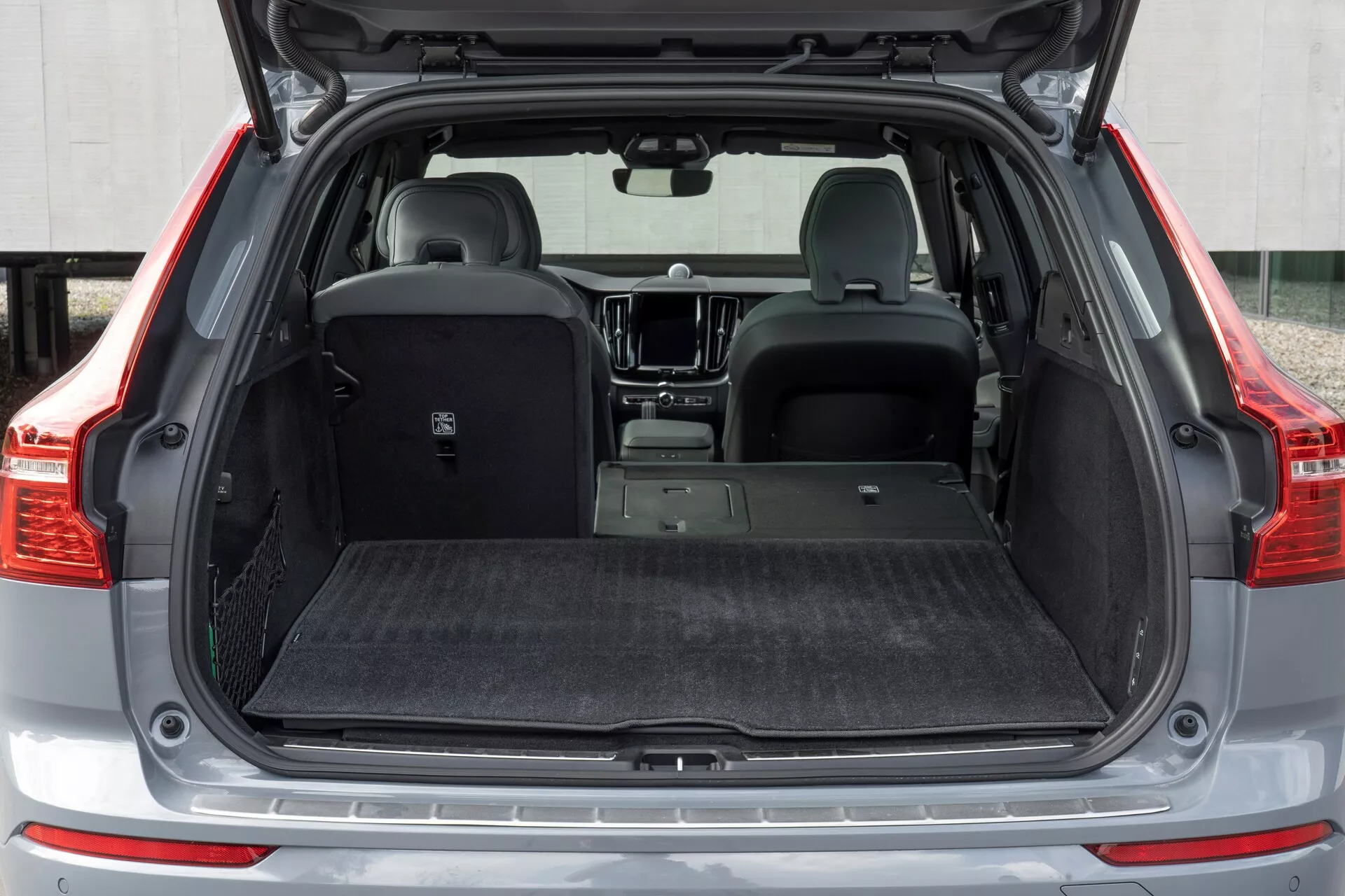 Närbild bakifrån av en 2021 Volvo XC60 andra generationen facelift, som visar den öppna bagageluckan från låg vinkel. Bilden framhäver det rymliga lastutrymmet med minimal hjulbågsinträngning, utdragbart lastskydd och interiört mattbelägg, inriktat på bullerreduktion och skydd. De delvis nedfällda baksätena visar på mångsidighet, medan ett skyddande lasttröskelskydd, synliga parkeringssensorer på den bakre stötfångaren, LED-belysning i bagageutrymmet och förankringspunkter betonar funktionalitet och detaljfokus.