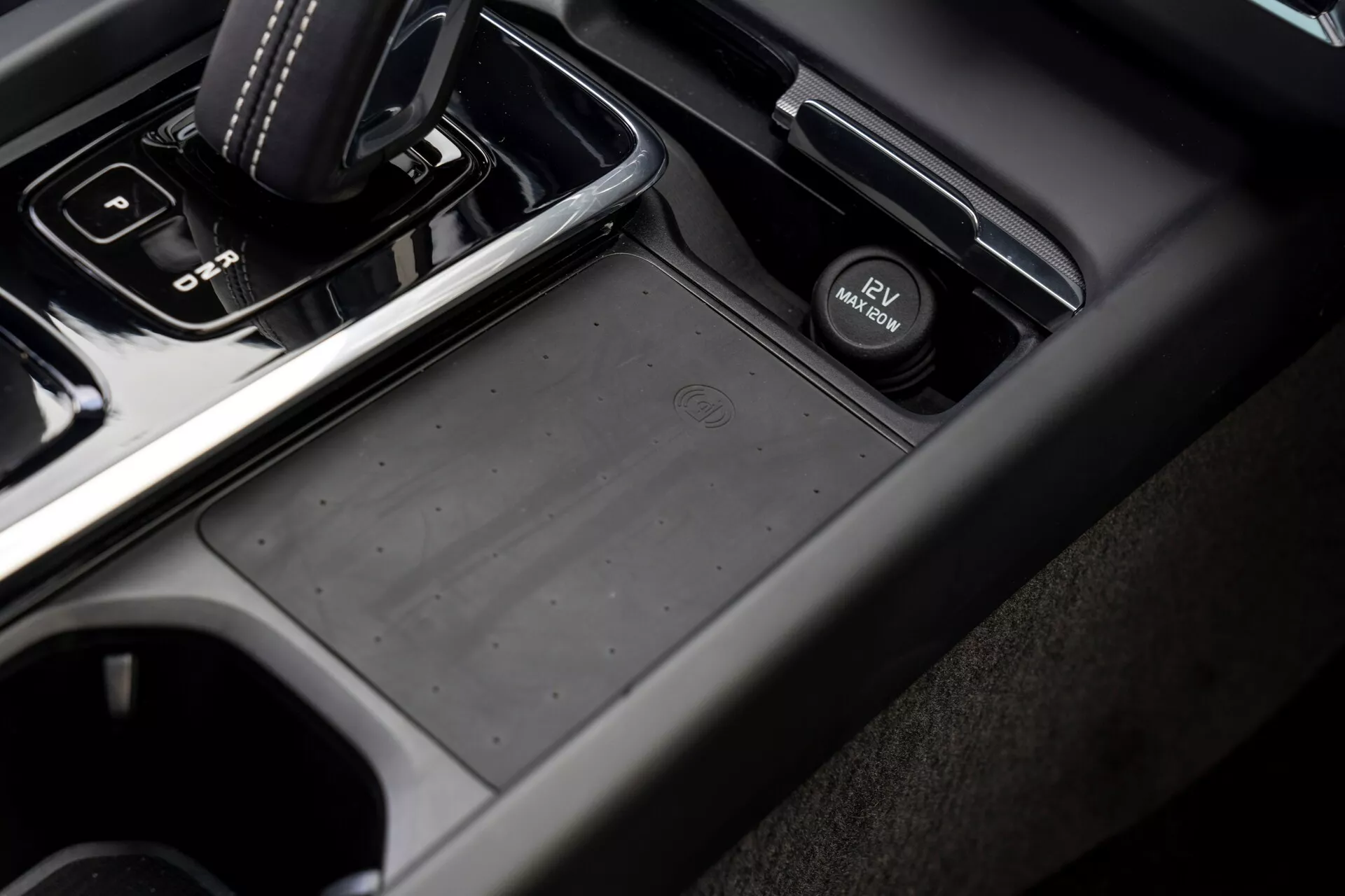 Närbild, vinklad vy av mittkonsolen i en Volvo XC60 2021 (andra generationen facelift) från passagerarsidan, med en automatisk växelspak i 'park' läge, ett halkfritt förvaringsfack, en knapp för elektronisk parkeringsbroms, kopphållare och en kombination av mjukplast och metallisk finish i dämpade mörkgrå och svarta toner.