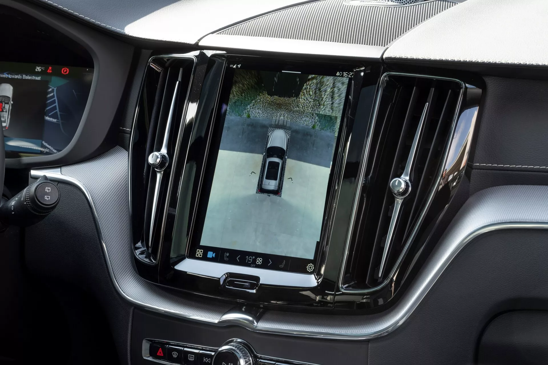 Interiörvy av en Volvo XC60 2021 (andra generationens facelift) som visar en stor, vertikalt orienterad infotainmentskärm på mittkonsolen och visar en bild från en backkamera med fågelperspektiv. Lyxig finish med kromdetaljer och eleganta designelement, ytterligare kontroller under skärmen och en delvy av ratten, vilket indikerar en premium och avancerad teknologisk interiör.