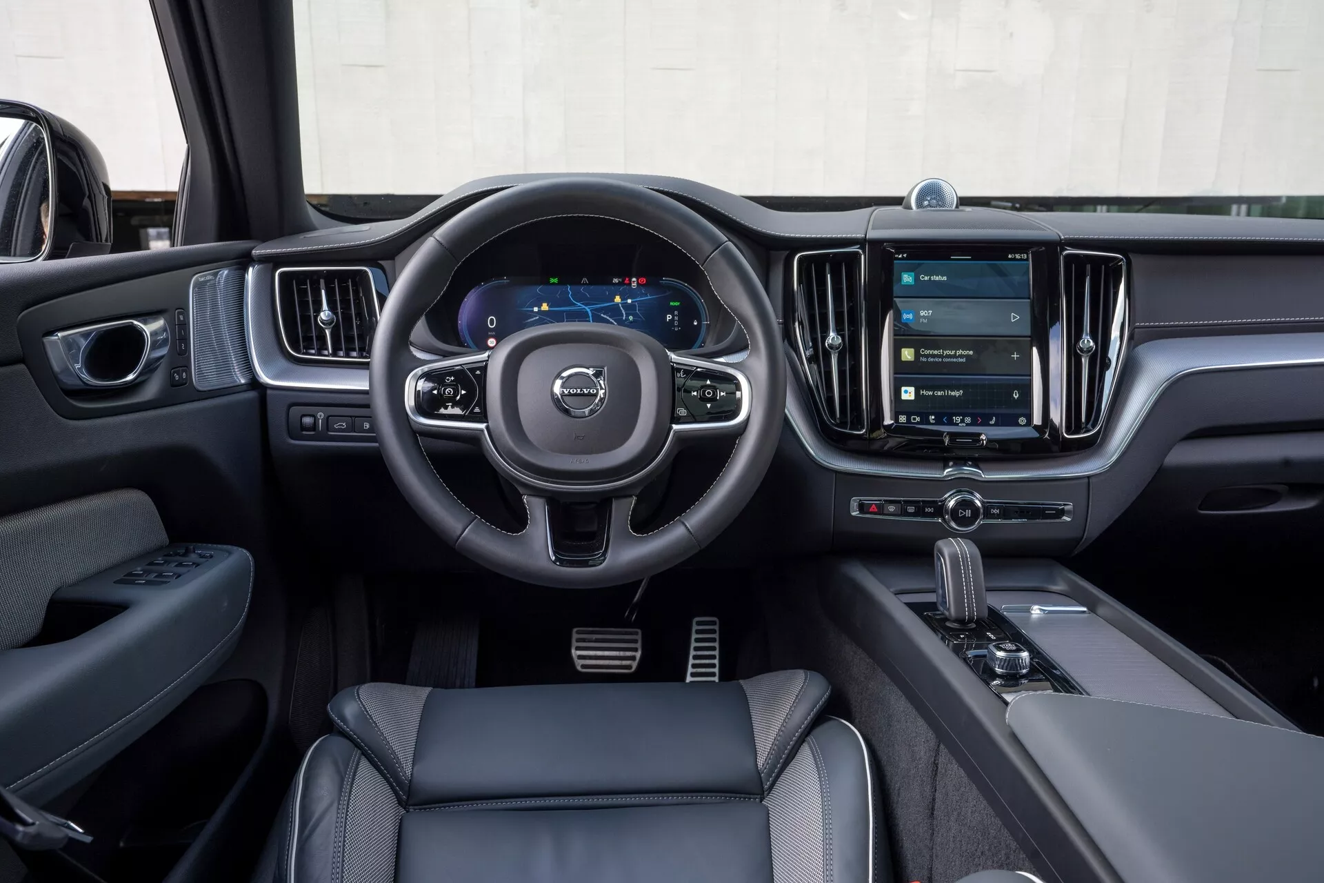 Interiör av en Volvo XC60 2021 (2:a generationens facelift) från förarens perspektiv. Bilden visar ratten med tre ekrar och kontrollknappar för ljud, kommunikation och farthållarinställningar, en digital instrumentpanel som visar hastighetsmätare och navigation, samt ett stort vertikalt pekskärmsinfotainmentsystem. Moderna designelement inkluderar läderliknande material, kromdetaljer och en automatisk växelspak med körlägesväljare. På förardörren syns fönster- och spegelreglage, vilket framhäver teknologiska och lyxiga egenskaper.