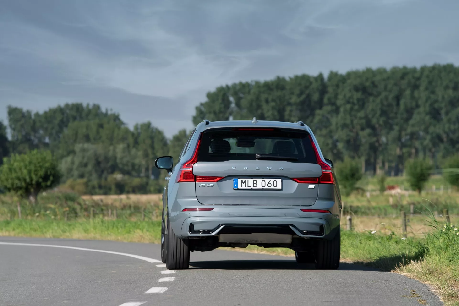 Bakifrån vy av en modern 2021 Volvo XC60 SUV (2:a generationen-facelift) på en tvåfilig väg i en naturlig, lantlig miljö, som visar distinkta bakljus, dubbla avgasrör, bakre spoiler med inbromsningsljus integrerat, backkamera, bakrutetorkare och parkeringssensorer. Placerad något till höger om mittlinjen, med klar himmel och träd i bakgrunden.