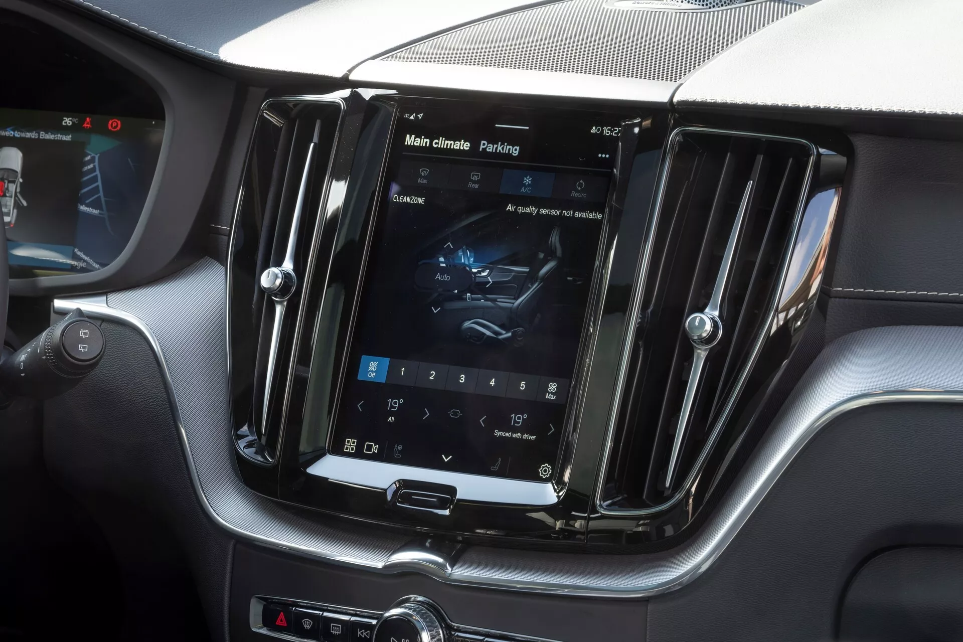 Interiörbild av 2021 Volvo XC60 (andra generationens ansiktslyft) med fokus på mittkonsolen. Bilden fångar den vertikalt orienterade infotainmentskärmen inuti en kromliknande ram, som visar klimatkontrollinställningar och en parkeringsassistansflik. En lyxig blandning av läder och kolfiberliknande trim omger skärmen, tillsammans med ett hybridstyrgränssnitt med fysiska knappar och vred. Fotot betonar avancerat infotainment, klimatkontroll, parkeringsassistans och lyxig interiördesign.
