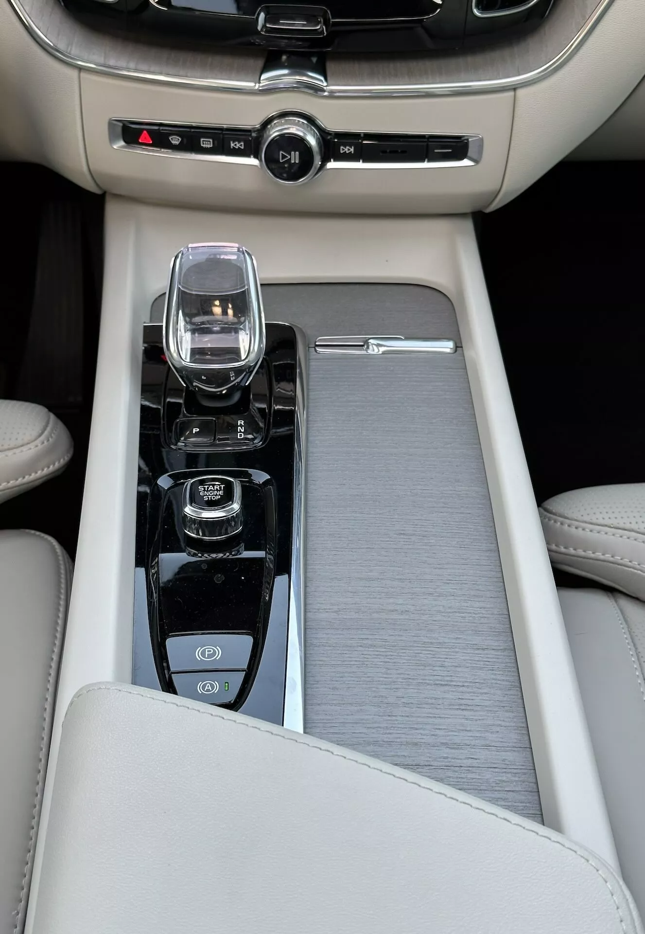 Närbild av mittkonsolen i en Volvo XC60 Recharge plug-in hybrid från 2021, med en beige läderarmstöd, ett skjutbart borstad metall- eller plastdekoration, en modern infotainmentkontroll, en kristallglas växelväljare för en automatisk växellåda, och kontroller för start/stopp-motorn, avstängning av dragkraftkontroll och kanske en lägesväljare för olika kördynamik, samt reglage för tvåzons klimatkontroll.