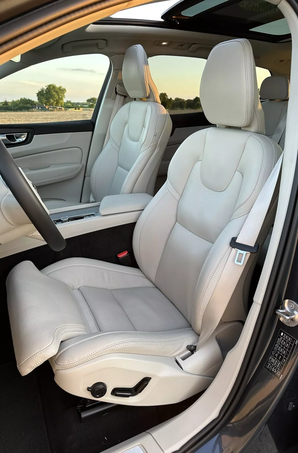 Interiörvy av Volvo XC60 Recharge plug-in hybrid 2021: fram- och delvis baksäten, lyxigt ljusfärgat läderklädsel, modern design, eljusterbara säten med eleganta sömmar, tagen från utsidan vid passagerarsidan dörr i dagsljus
