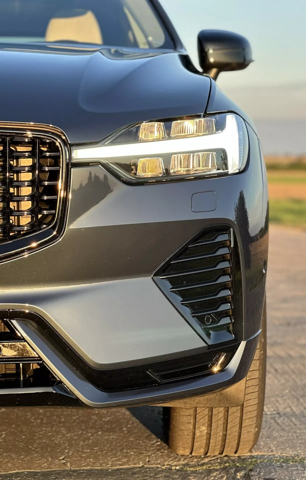 Närbild på främre kvartalet av en Volvo XC60 Recharge Plug-In Hybrid 2021, som framhäver dess vertikala grill, LED-strålkastare med dagljuslampor, stylade ventiler och dimljus, sportig huva och skärmar, metallisk lack med kontrasterande trim, och en glimt av dess robusta hjul och däck, förhöjd av fordonets glansiga finish.