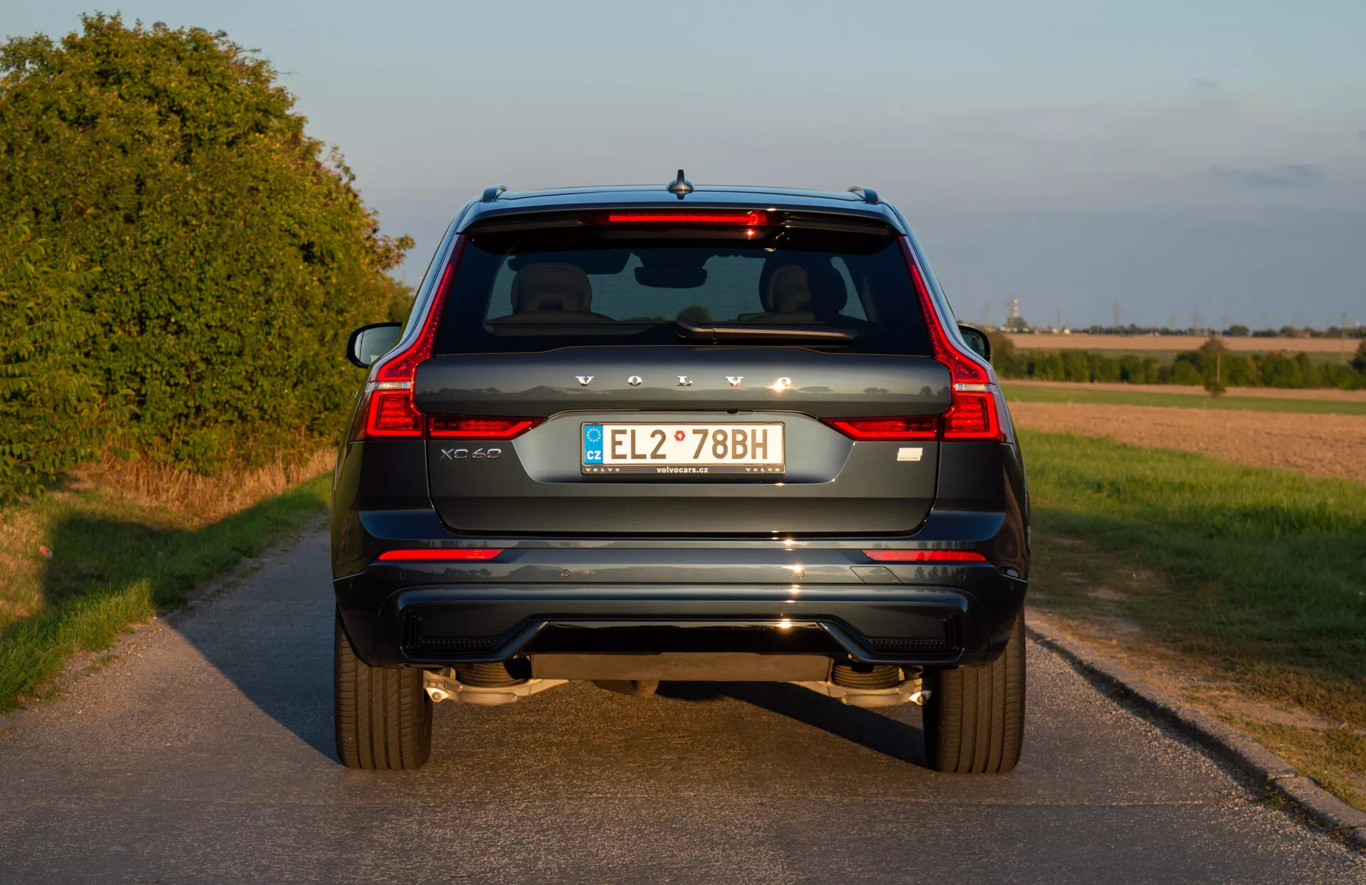 Bakifrån vy av en blå 2021 Volvo XC60 Recharge Plug-In Hybrid SUV med vertikala bakljus, dubbla avgastips, bakre reflektorer, bakrutetorkare, tredje bromsljus, europeisk registreringsskylt och backkamera på en landsväg före solnedgången