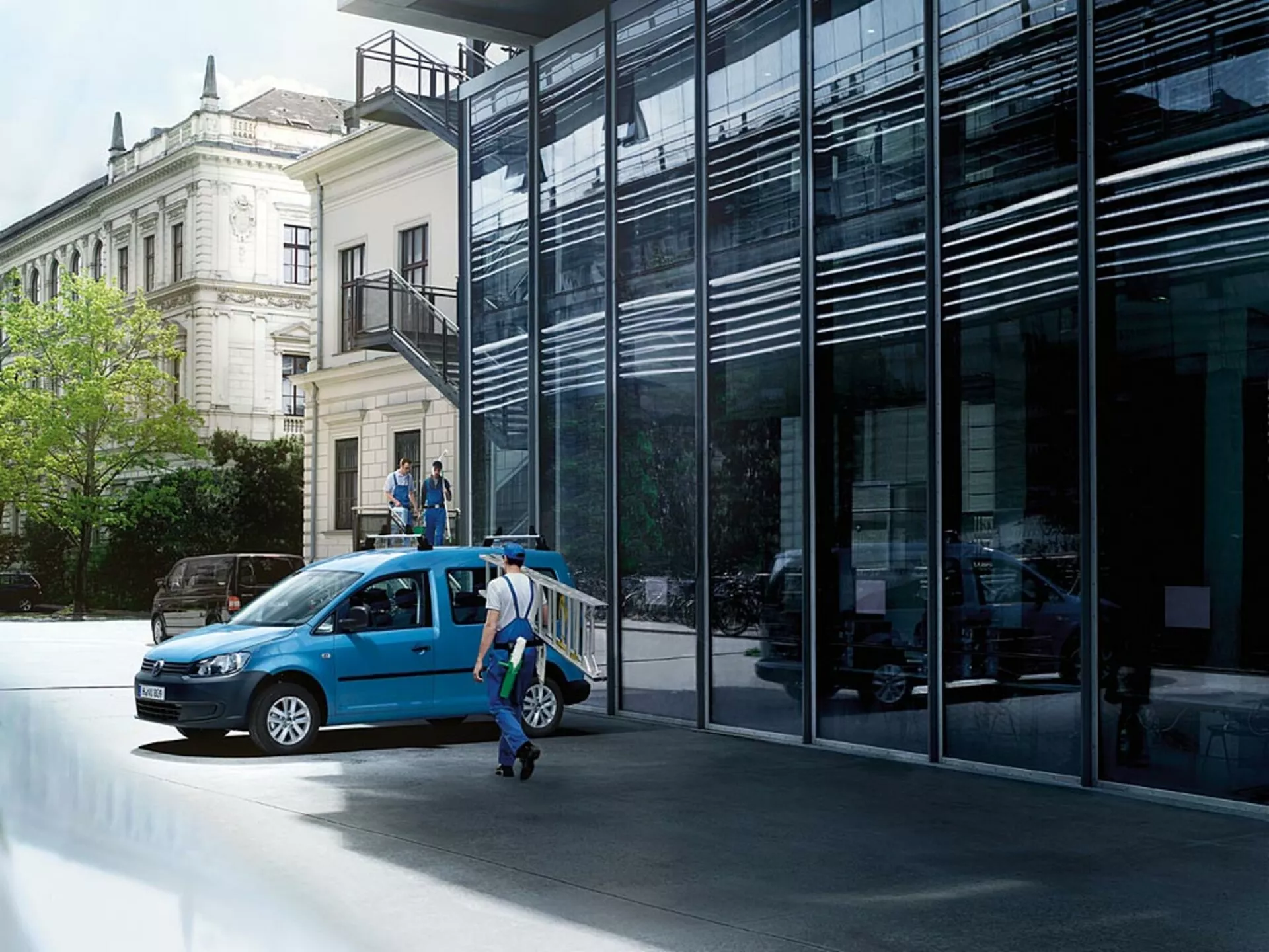Blå VW Caddy från 2010 parkerad på en stadsgata, visas från en trekvarts framifrån vy med öppna dörrar och betonar dess frontgrill, strålkastare, hjul och en glimt av interiören mot en bakgrund av modern och klassisk arkitektur