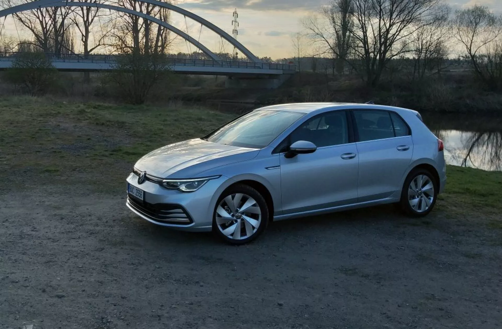 Volkswagen Golf GTD från 2017 i silver, visad från ett främre trekvarts perspektiv som belyser dess eleganta horisontella LED-strålkastare, minimalistisk stängd grill, distinkta lättmetallhjul och aerodynamiska karosslinjer, parkerad på en oasfalterad yta med lugnt vatten och en bågbro i bakgrunden under gyllene timmen