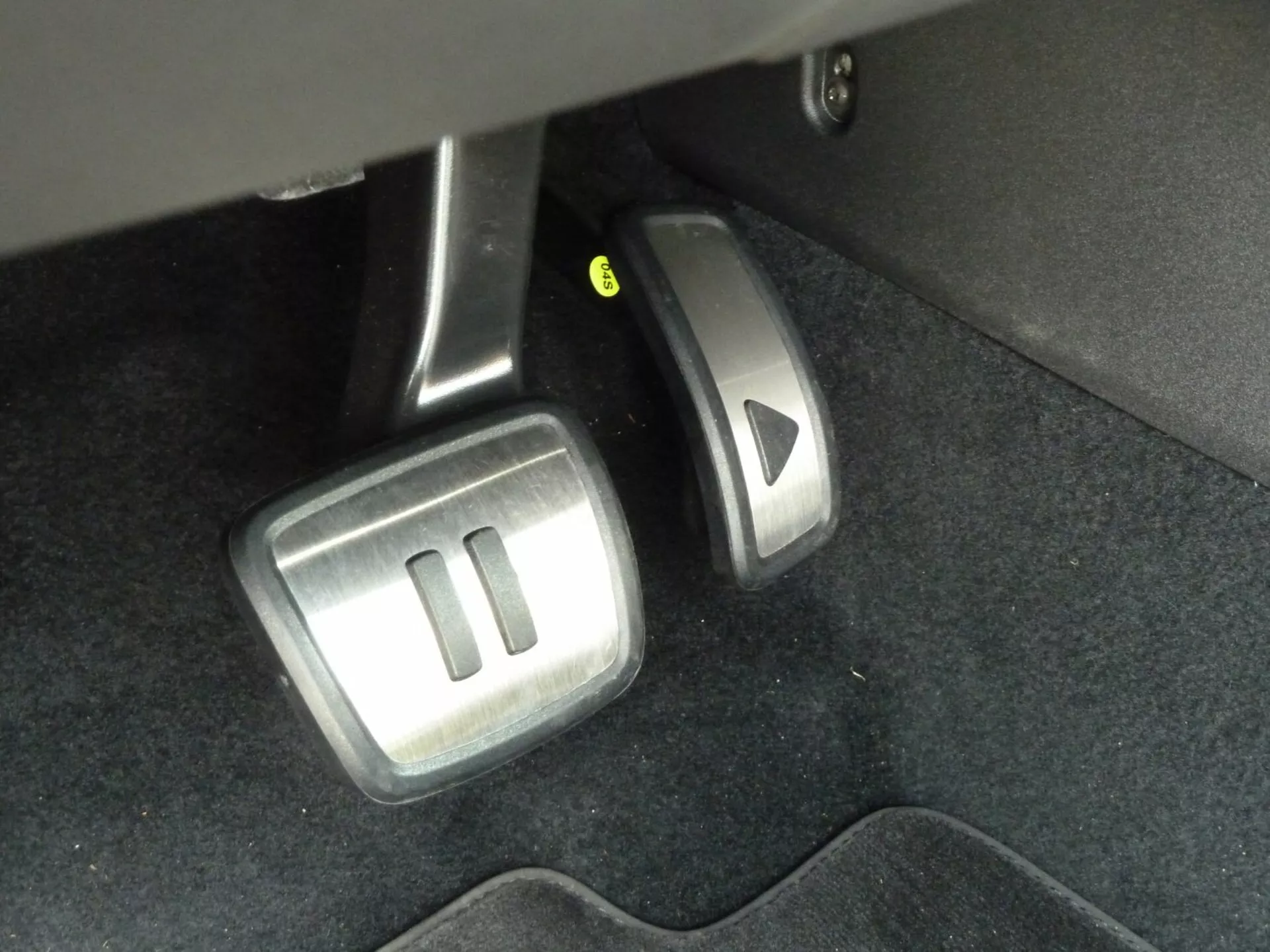 Närbild av VW ID3 2020 manuell växellåda bilpedaler från förarens perspektiv, som visar kopplings-, broms- och gaspedaler med metalliska och gummigrepp ytor, tillsammans med en potentiell styrkolumnsjusteringshävstång