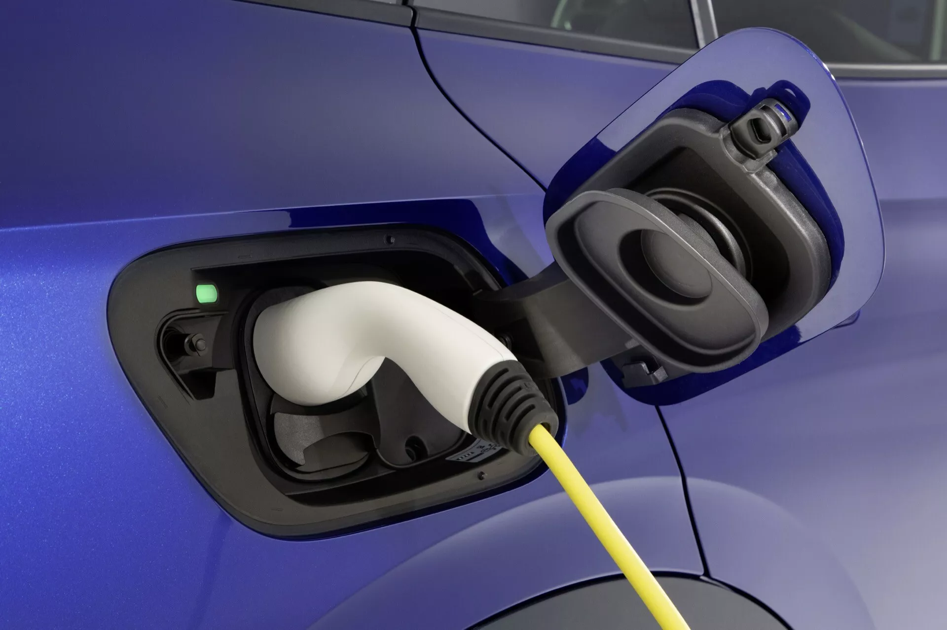 Närbild av VW ID.4 2020 (1:a generationen) elektriska fordonets laddningsportområde med öppet lock som visar uttaget och den vita kontakten inpluggad, markerad av en grön laddningsindikatorlampa, mot en blå bilkropp.