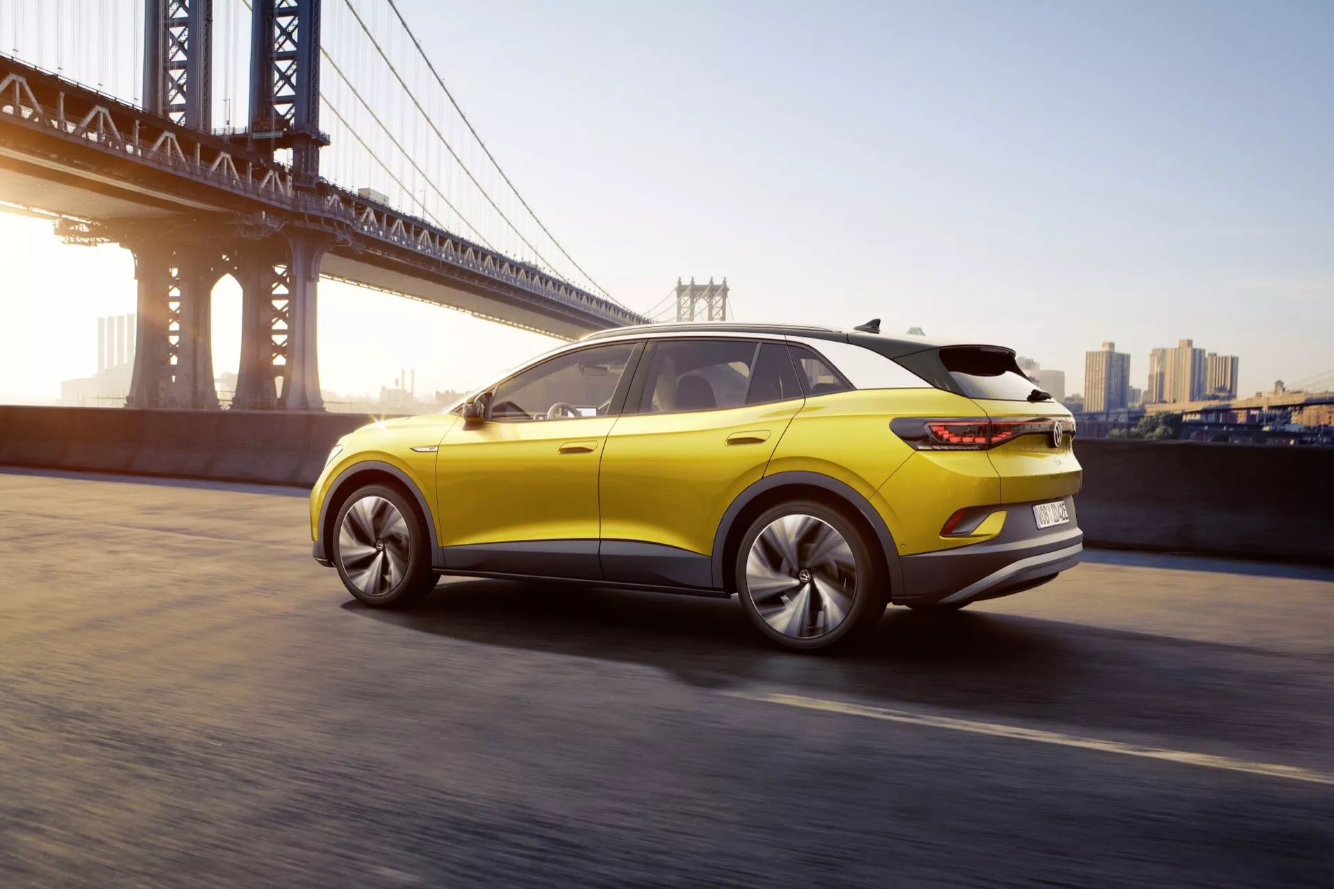 Dynamisk bakre trekvarts vy av en gul VW ID.4 från 2020, som framhäver dess sportiga sluttande taklinje, svepande LED-bakljus och flerspakiga legeringshjul, mot en urban bakgrund med en hängbro och stadssiluett i gryningen eller skymningen.