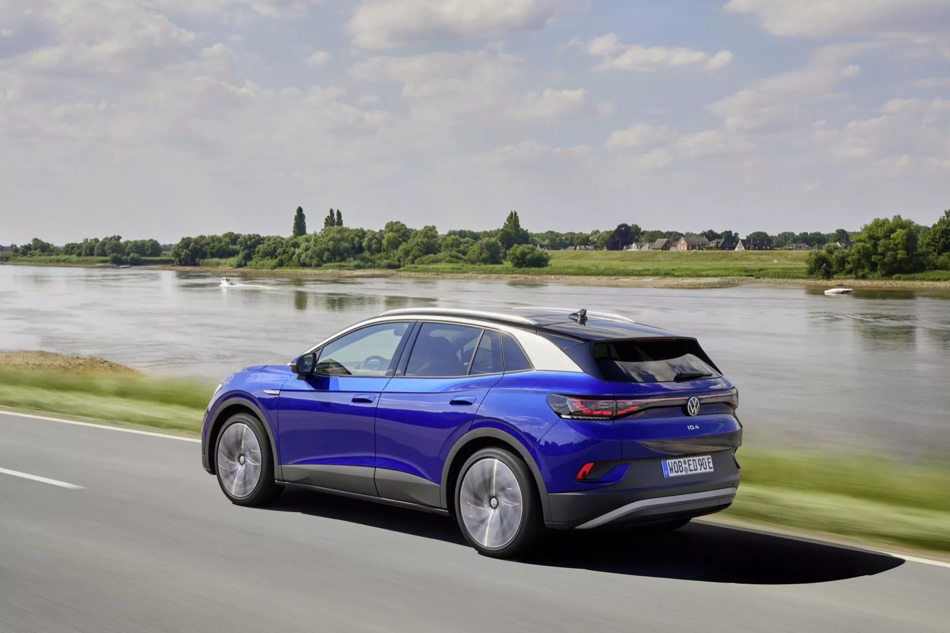Dynamisk sidoprofil av VW ID.4 1:a generationen 2020 i livligt blått, med sluttande taklinje, sportig spoiler, flerspakiga lättmetallfälgar och avancerad LED-teknik, som förmedlar rörelse på en väg vid en flod