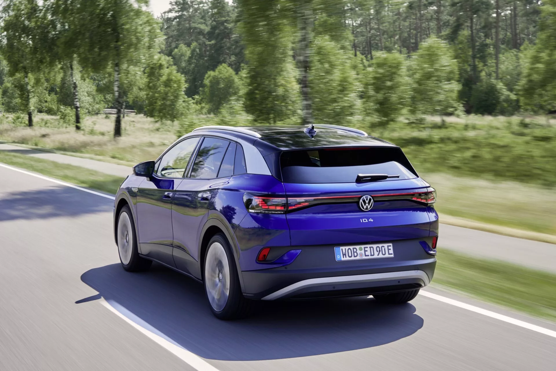 2020 Volkswagen ID.3 hatchback i rörelse på en solig väg, visas från ett bakre trekvarts perspektiv som framhäver dess eleganta blåa lack, kontinuerligt LED-bakljusbälte, aerodynamiska hjul och smarta teknikfunktioner inklusive sensorassisterad körning och elektrisk effektivitet.