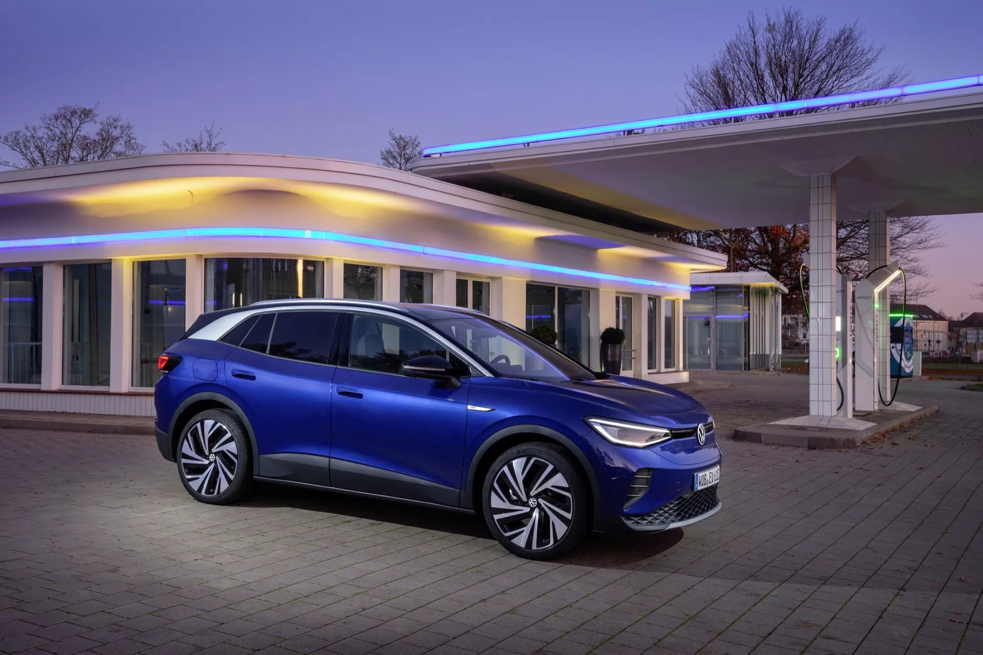 VW ID.4 första generationen från 2020 parkerad utomhus i skymningen, som visar dess metalliska blå sida profil mot en modern byggnad med varm belysning, vilket framhäver SUV:ens eleganta design, LED-belysning och avancerad aerodynamik.