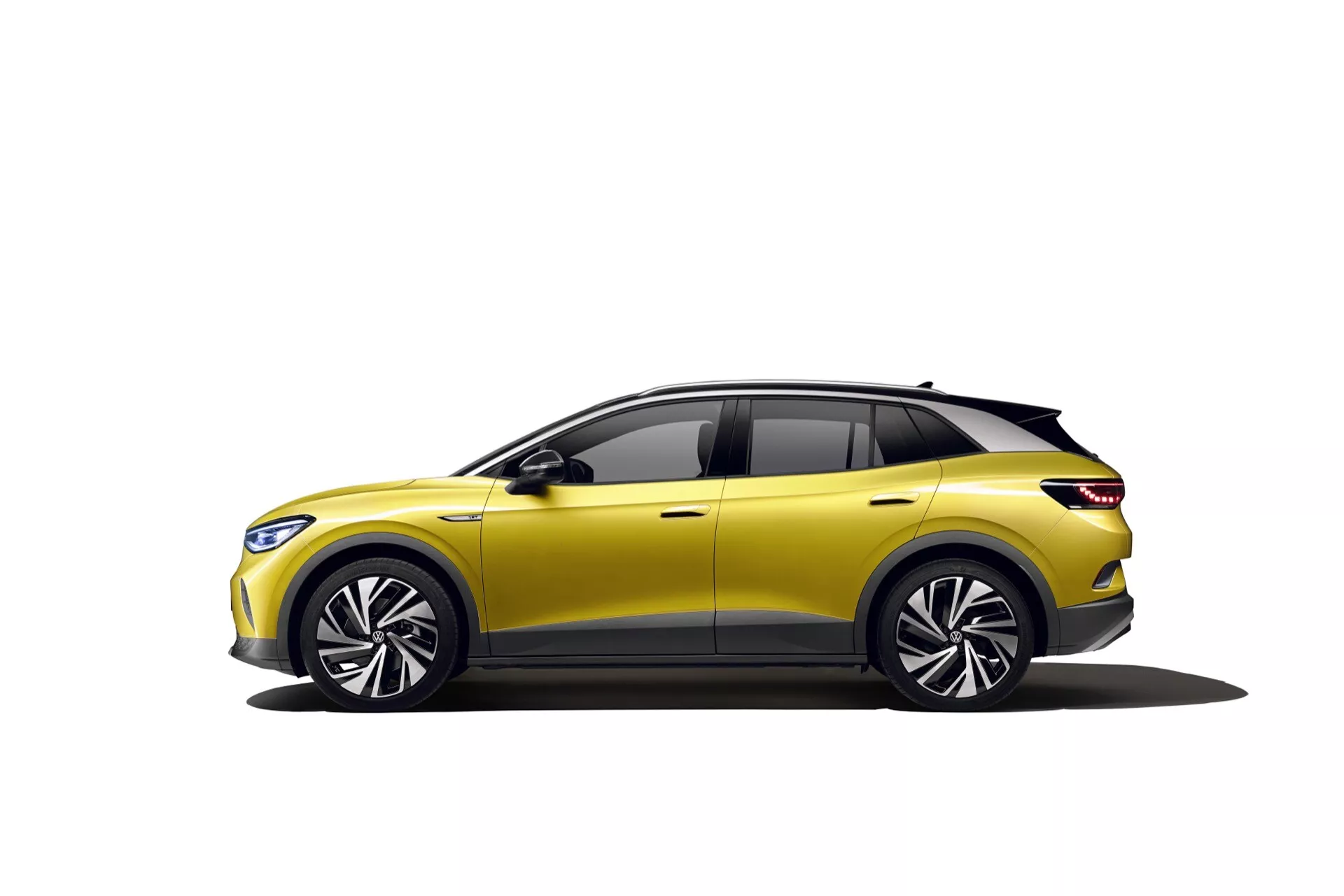 VW ID.4 första generation 2020 i gult, visar upp sin moderna crossover-design, femdörrars kaross, stora flerspakiga lättmetallhjul, dynamisk sidoprofil med karaktärslinje, integrerade blinkers i sidospeglarna, fönster med svarta kanter och tonade rutor, aerodynamiskt sluttande tak med spoiler och panoramataket, delvis synliga eleganta främre strålkastare, mot en vit bakgrund