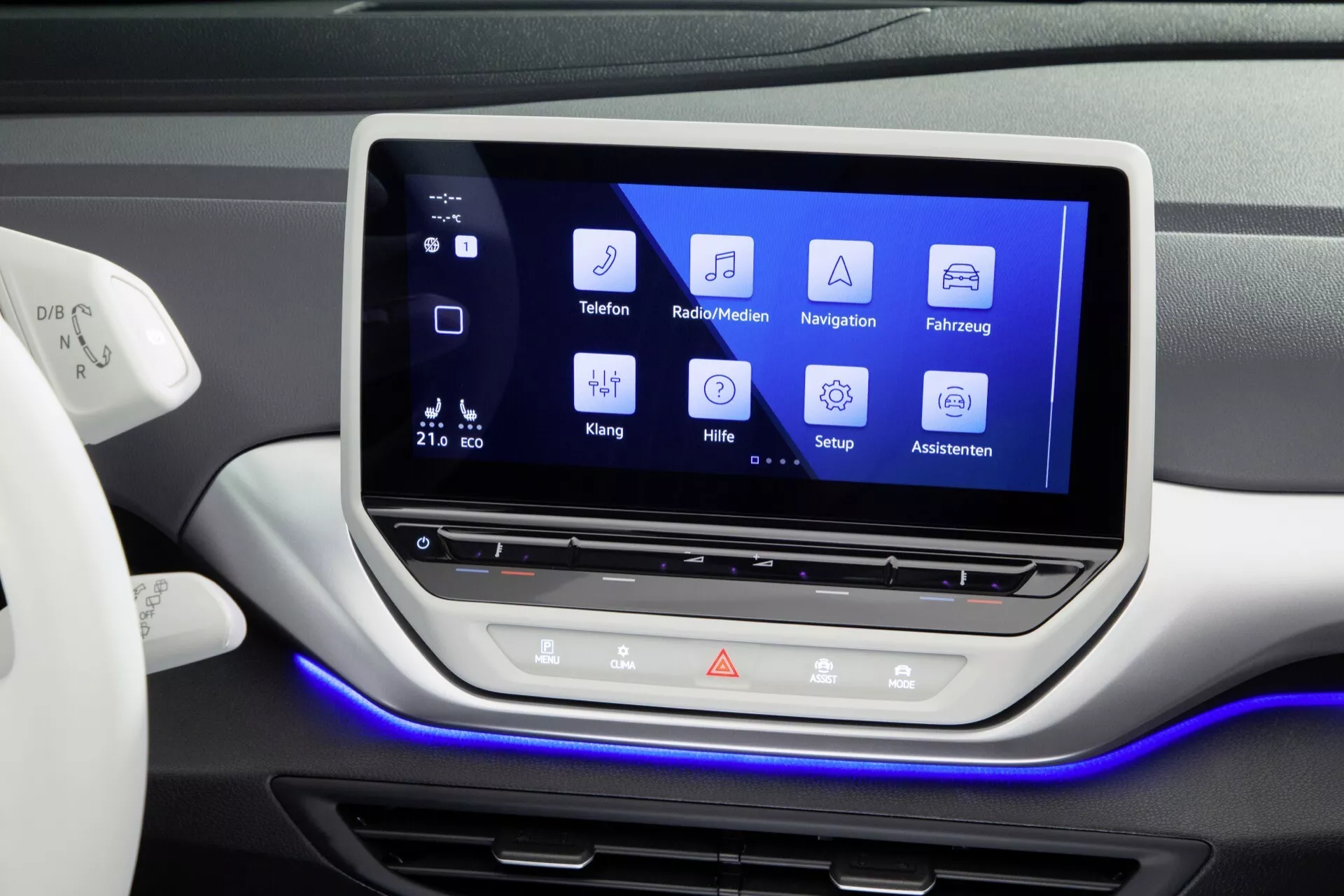 Centrala instrumentbrädan på en VW ID.4 2020 med ett stort pekskärmsinfotainmentsystem med ikoner för Telefon, Radio/Media, Navigation, Fordonsinställningar, Ljudinställningar, Hjälp, Inställningar och Assistent; utrustad med fysiska knappar och central knapp under skärmen, omgivningsljus och indikation på klimatkontroll