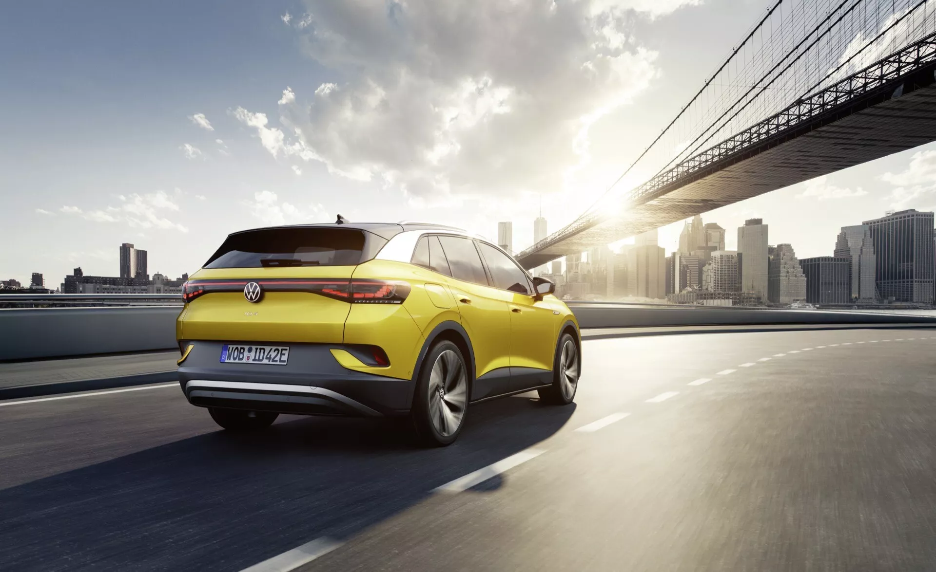 VW ID.4 1:a generationen från 2020 i gult körande på en bro vid solnedgången, visar bak- och sidoprofil med LED-bakljus, aerodynamisk spoiler och lättmetallhjul