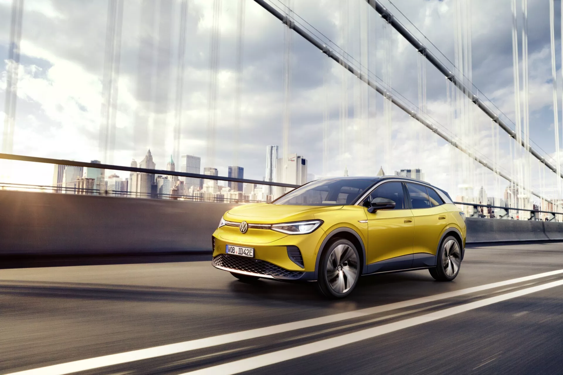 VW ID.4 1:a generationen 2020 i rörelse, visar dynamisk design och tekniska funktioner från ett lågt vinkel framifrån vänster trekvarts vy, med fordonets iögonfallande gula färg, moderna legeringshjul och potentiella ADAS framhävda mot en suddig stadsbakgrund