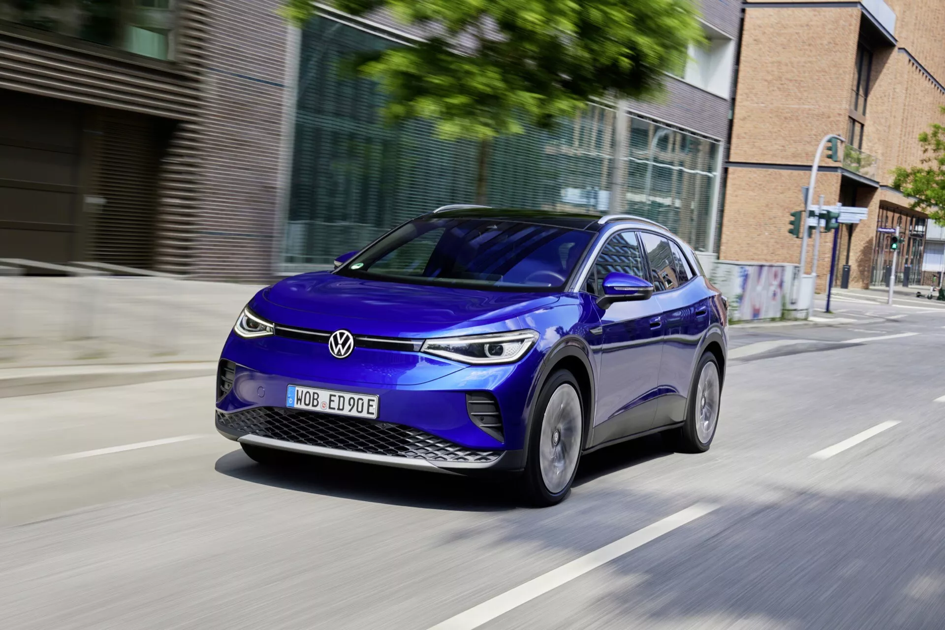 Dynamisk bild av en blå fyradörrars halvkombi-stil elektrisk bil VW ID.4 1:a generation 2020 i rörelse på en stadsväg, visar en front tre fjärdedels vy som framhäver modern design, LED-strålkastare, dagtid körningsljus och aerodynamiska legeringsfälgar
