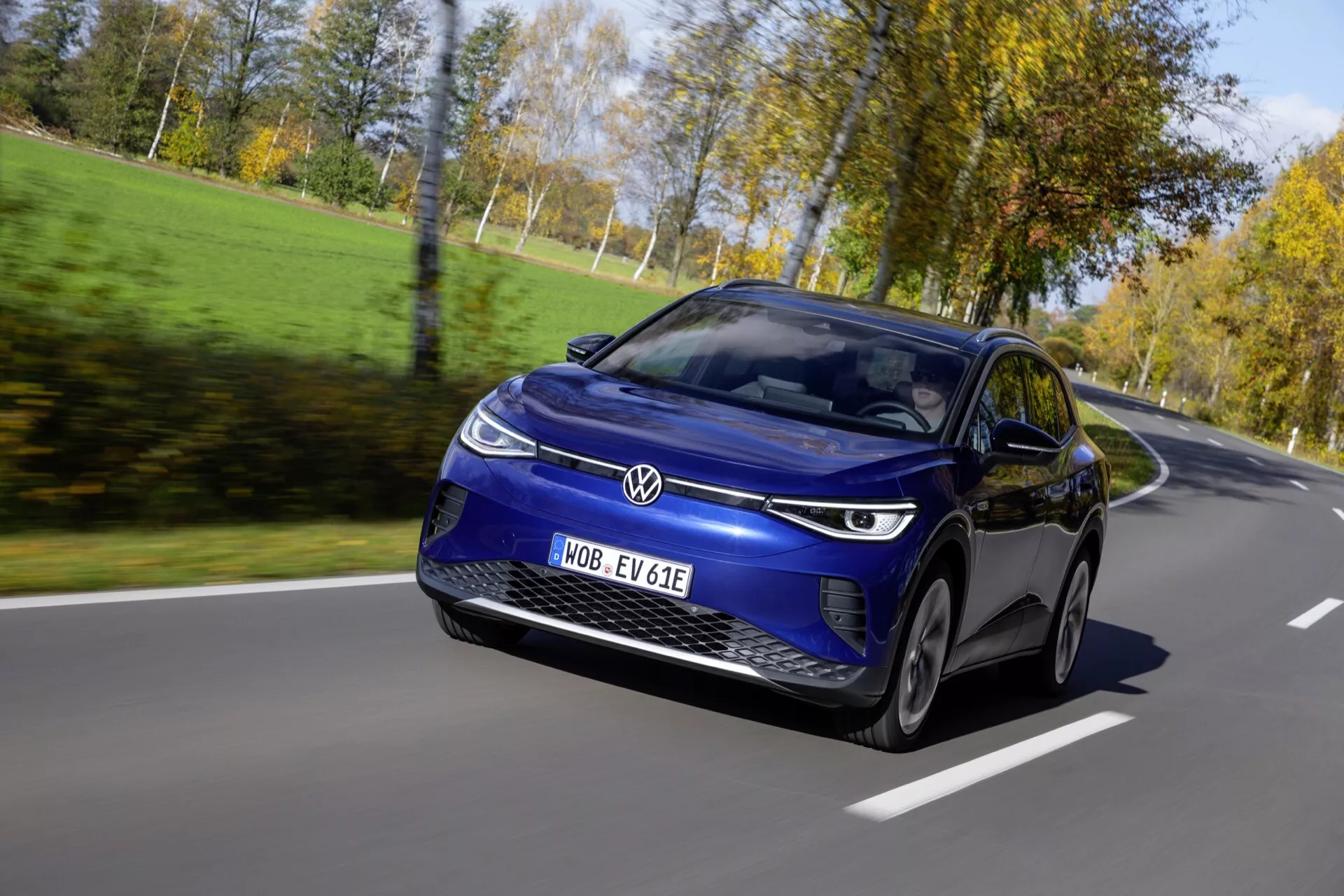 Dynamisk främre tre fjärdedels vy av Volkswagen ID.4 1:a Generationen 2020 i rörelse på en klar dag, visar dess aerodynamiska blå hatchbackdesign, distinkta LED-strålkastare och avancerade teknikfunktioner inklusive digital display och märkning för elektriskt fordon