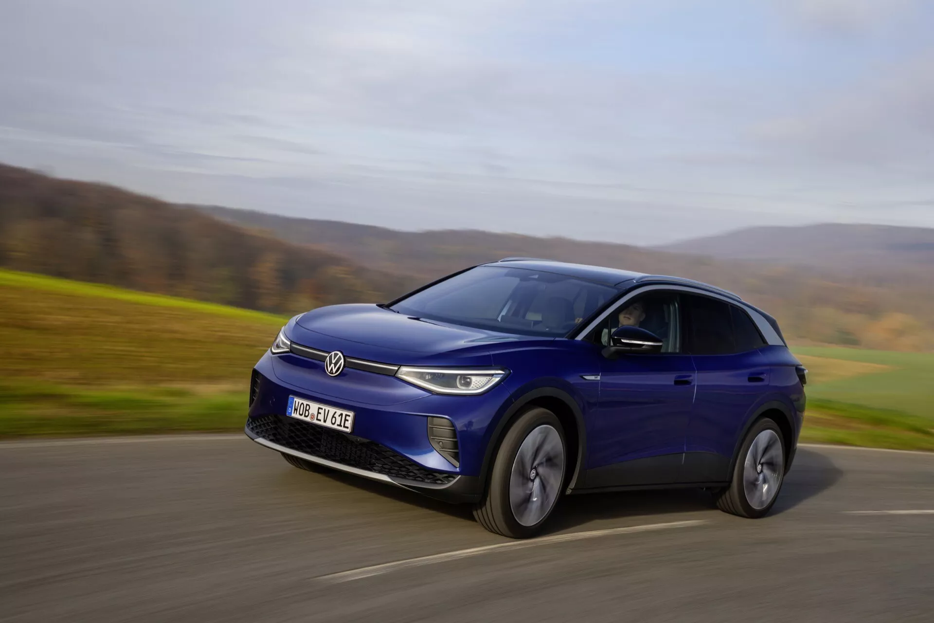 Blå Volkswagen ID.4 elektrisk crossover SUV i rörelse på en landsväg, fångad från ett främre tre kvarts perspektiv som belyser dess dynamiska rörelse och designfunktioner såsom den slutna grillen med LED-strålkastare, legeringshjul och en slank, sportig silhuett under en metallisk blå finish.