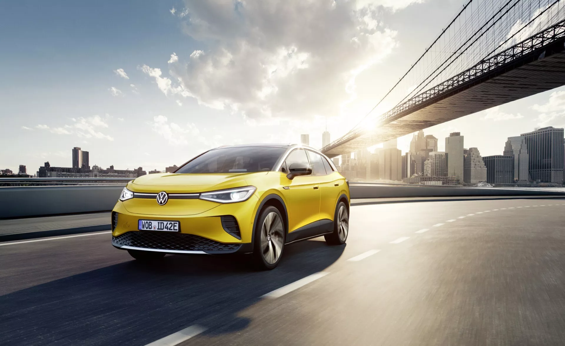 Dynamisk bild av en ljusgul Volkswagen ID4 2020 elbil i rörelse på en väg, som visar dess sportiga profil och moderna designfunktioner som LED-strålkastare, aerodynamisk kaross och legeringshjul mot en bakgrund av en stads skyline vid soluppgång eller solnedgång.