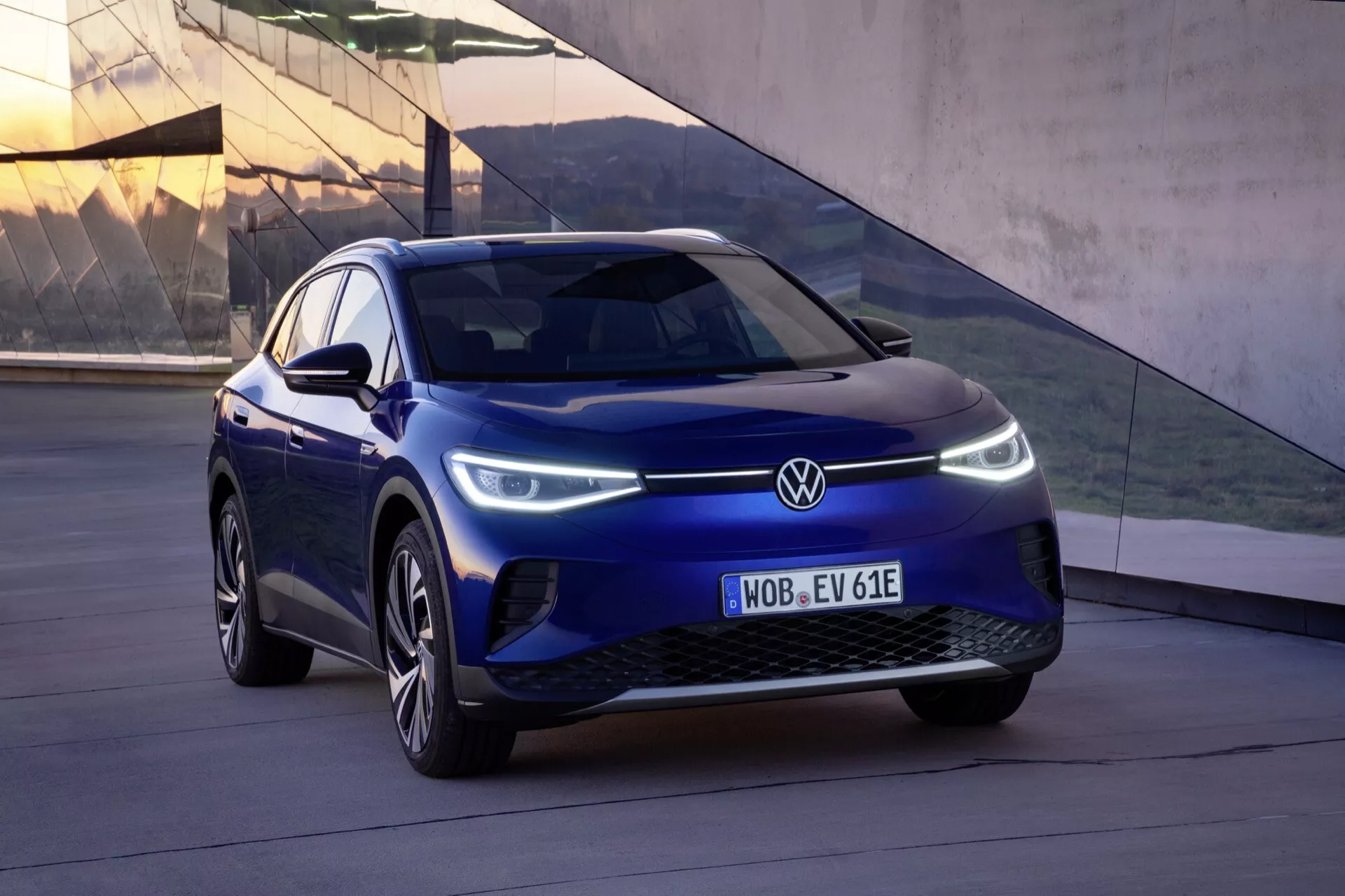 2020 VW ID.4 första generationen i blått, fotograferad från en låg vinkel framifrån tre fjärdedelar som visar fordonets vänstra sida, modern stängd grill, tillverkarens emblem, slanka LED-strålkastare med dagljusljus, aerodynamiska legeringshjul, integrerade blinkers på sidospeglarna, förarsidans laddningslucka, och skulpterad kaross för förbättrad aerodynamik och stil.