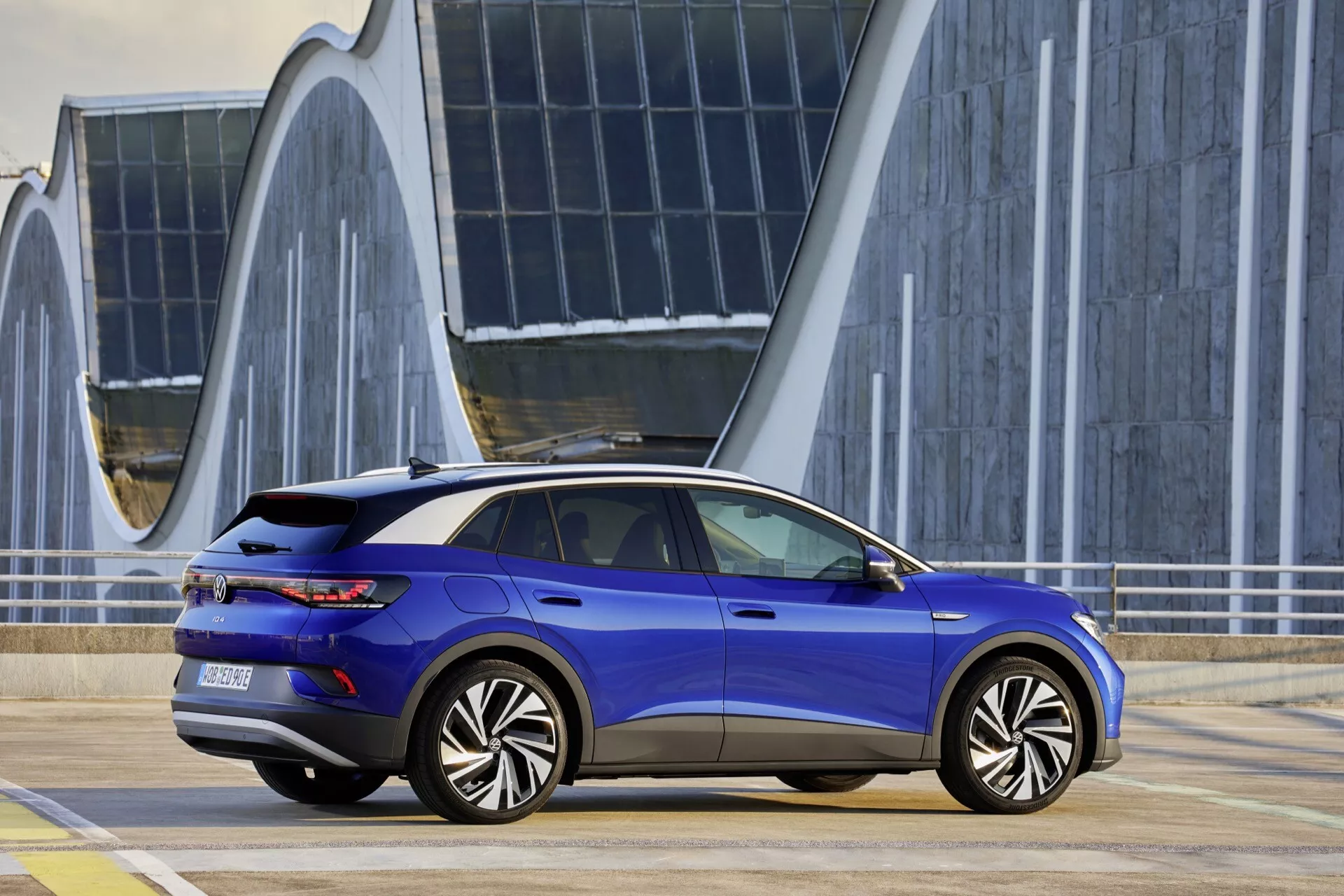 VW ID.4 1:a generationen 2020 i metalliskt blått, visar en stilig sida och bakprofil med aerodynamisk design, dörrhandtag i linje med karossen och integrerat ljusfält, parkerad på ett platt område med modern arkitektur i bakgrunden