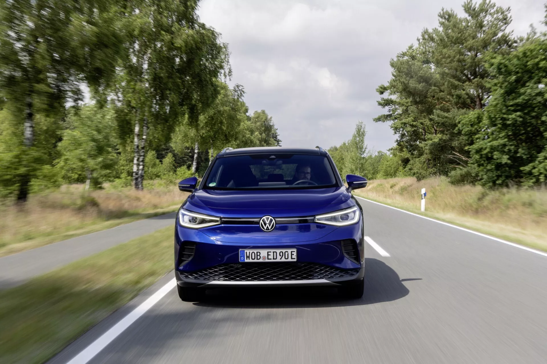 Frontal perspektiv av en blå VW ID4 2020 som kör på en väg omgiven av grönska, belyser dess moderna elbilsdesign, aerodynamiska linjer och avancerade LED-strålkastare.