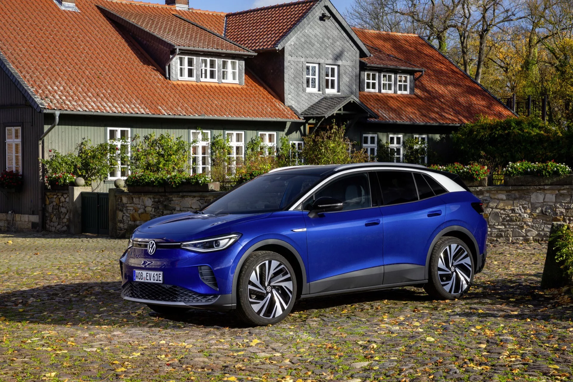 VW ID.4 1:a Generation 2020 parkerad på kullersten med höstlöv, framhäver dess moderna blåa elektriska SUV-design, avancerade LED-strålkastare, aerodynamiska hjul och potentiella förarassistanssystem i en fridfull historisk miljö.