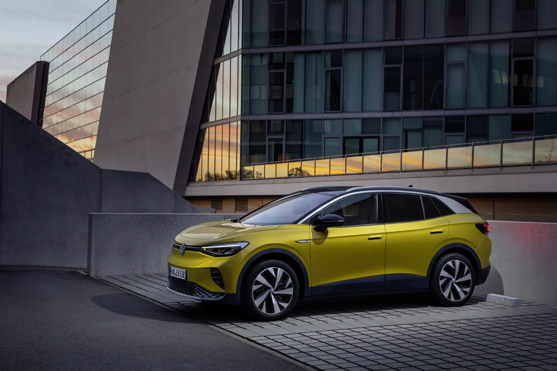 VW ID.4 1:a generationen från 2020 i lysande gult parkerad utanför en samtida byggnad, visar slanka LED-strålkastare, minimal grill som indikerar elektrisk eller hybrid design, skulpterad aerodynamisk huva, stora legeringshjul med flerspakad design, integrerade blinkers i sidobackspeglarna, distinkt karaktärslinje, svart fönstertrim med soltak, och unik design på bakdörrsfönstret som speglar modern fordonteknik och premiumfunktioner mot en lugn stadsbakgrund.